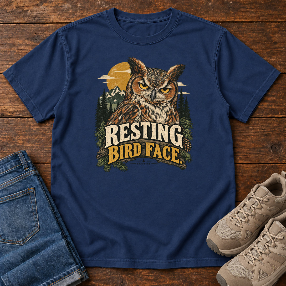 True Navy Resting Bird Face T-Shirt with a yellow-eyed, tufted owl with pine branches, mountains and a golden sun; front flat lay on wood.