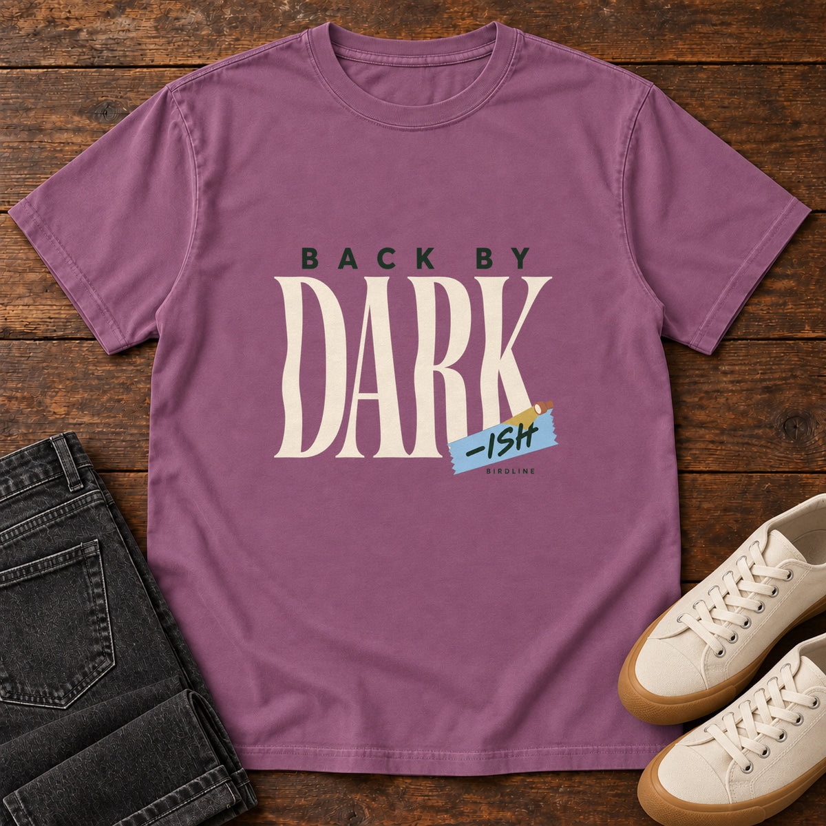 Berry Back by Dark-ish T-Shirt with tall cream Dark lettering and a small blue -ish label, front flat lay on wood.