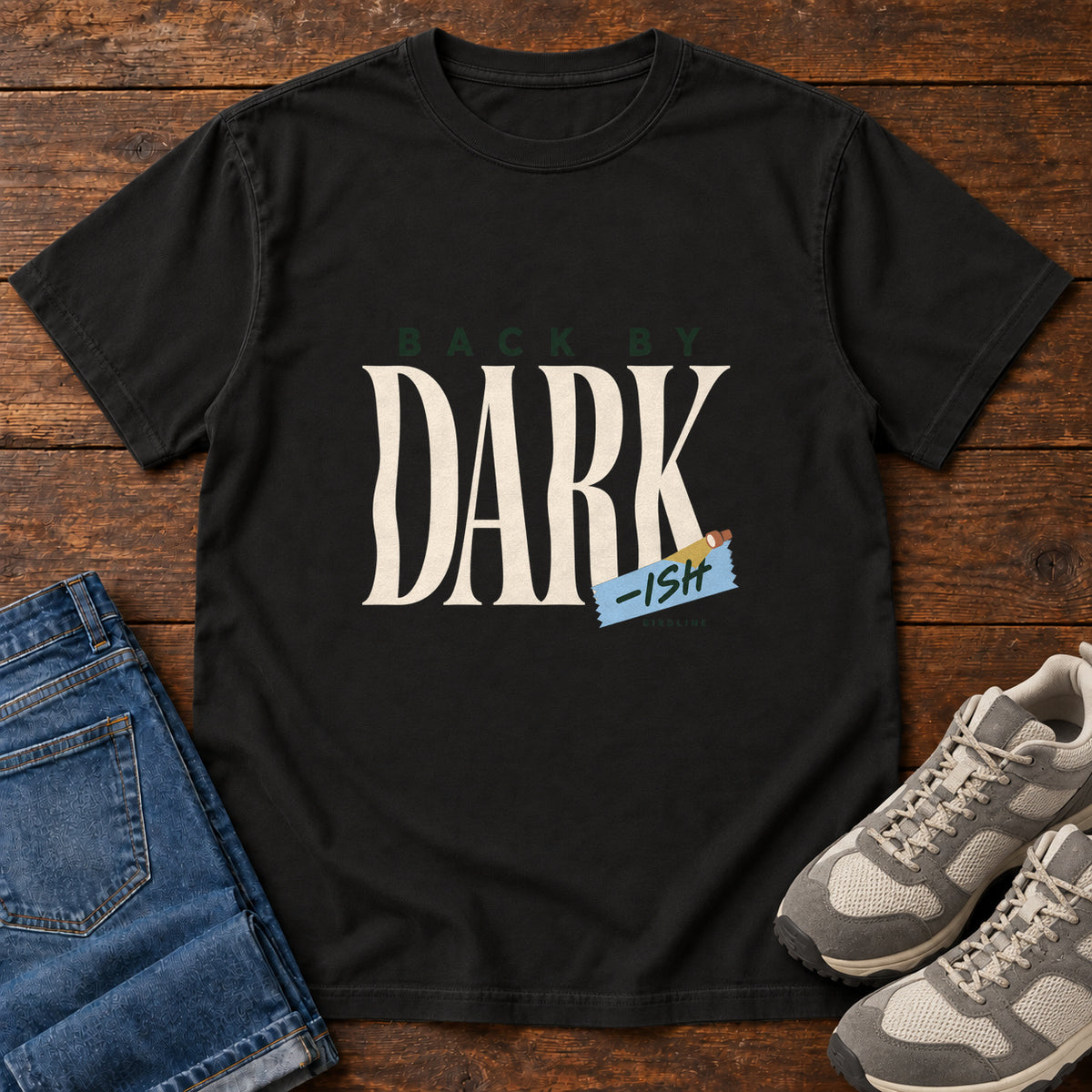 Black Back by Dark-ish T-Shirt with tall cream Dark lettering and a small blue -ish label, front flat lay on wood.