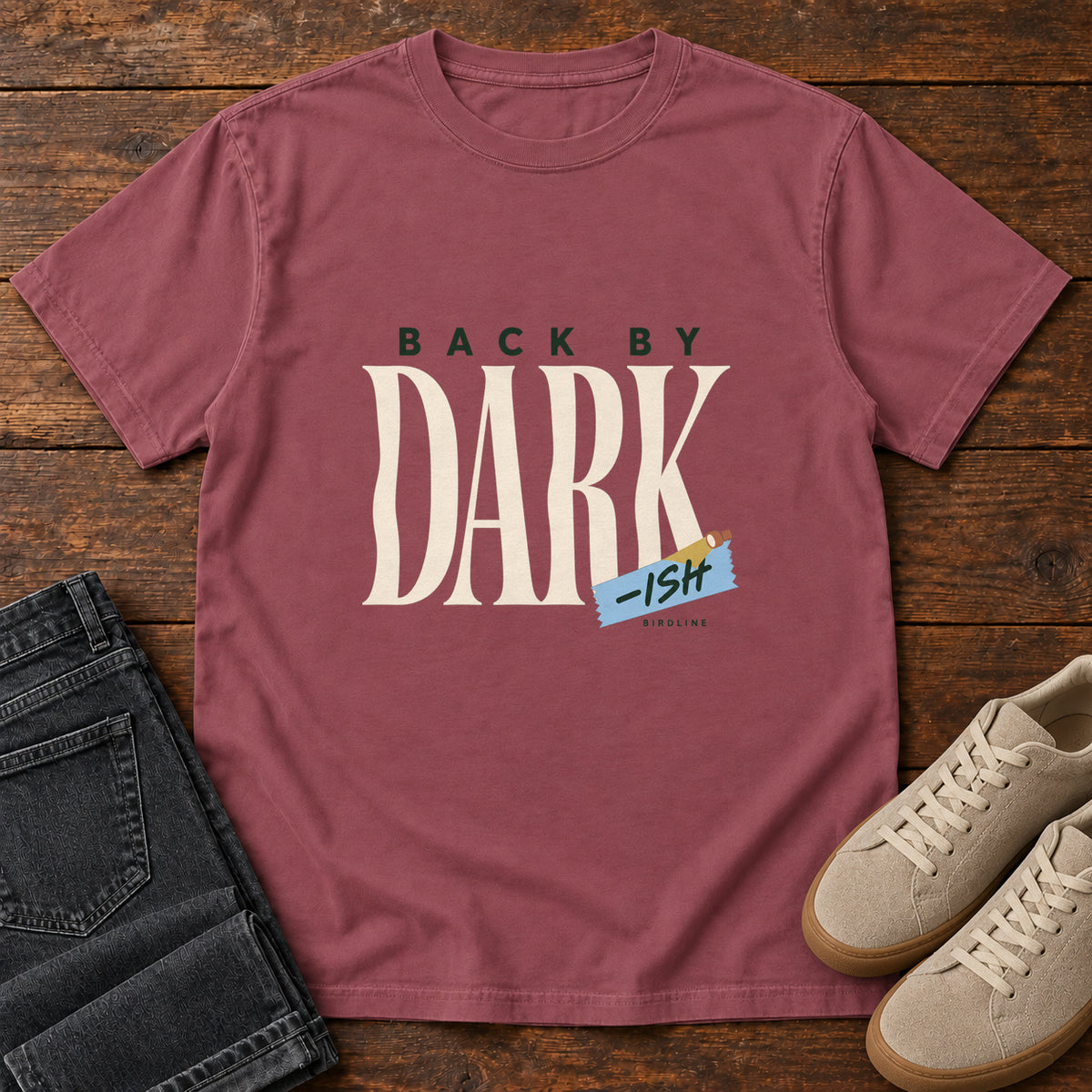 Brick Back by Dark-ish T-Shirt with tall cream Dark lettering and a small blue -ish label, front flat lay on wood.