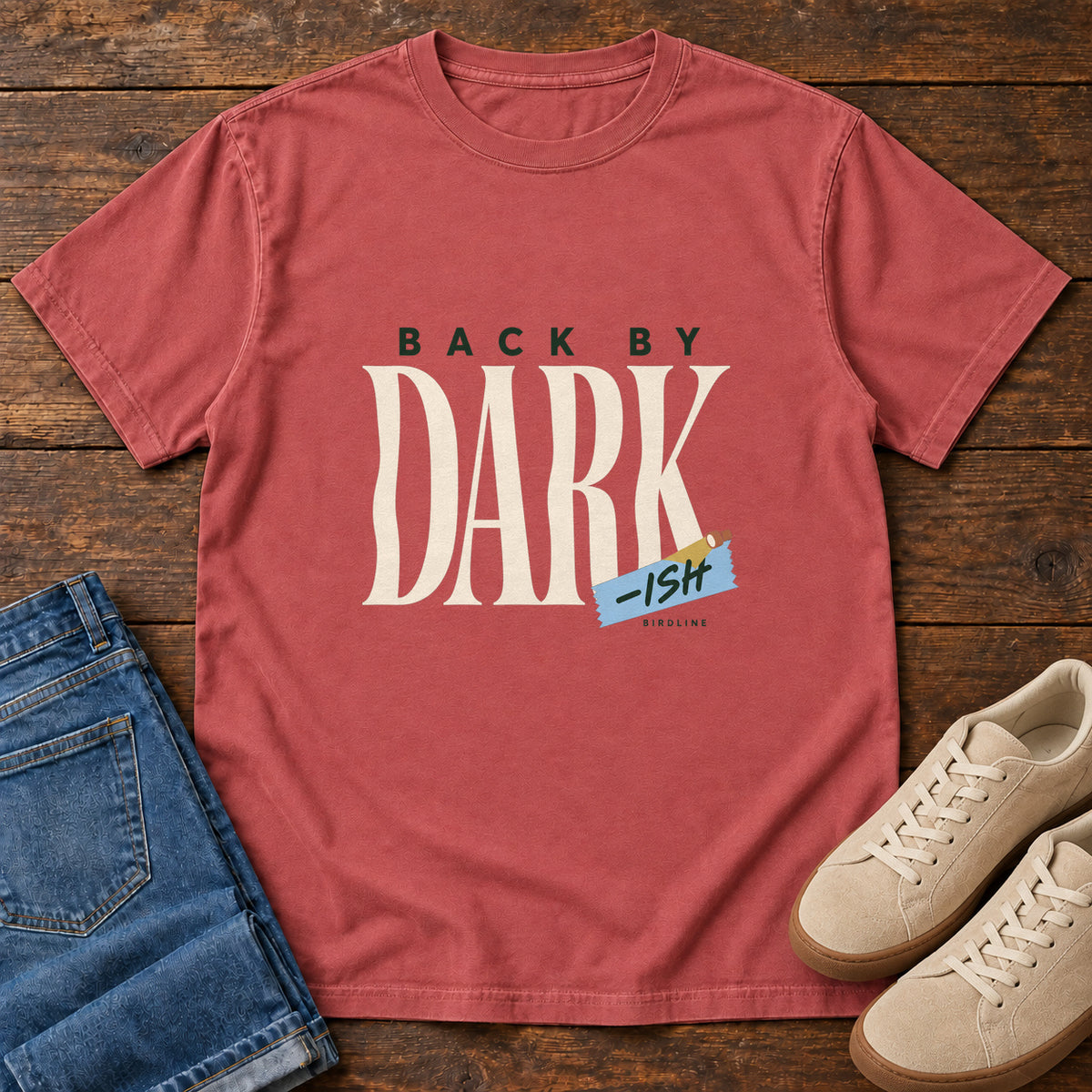 Crimson Back by Dark-ish T-Shirt with tall cream Dark lettering and a small blue -ish label, front flat lay on wood.