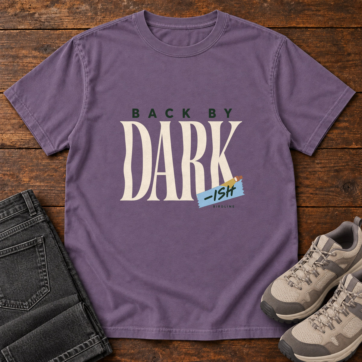 Grape Back by Dark-ish T-Shirt with tall cream Dark lettering and a small blue -ish label, front flat lay on wood.