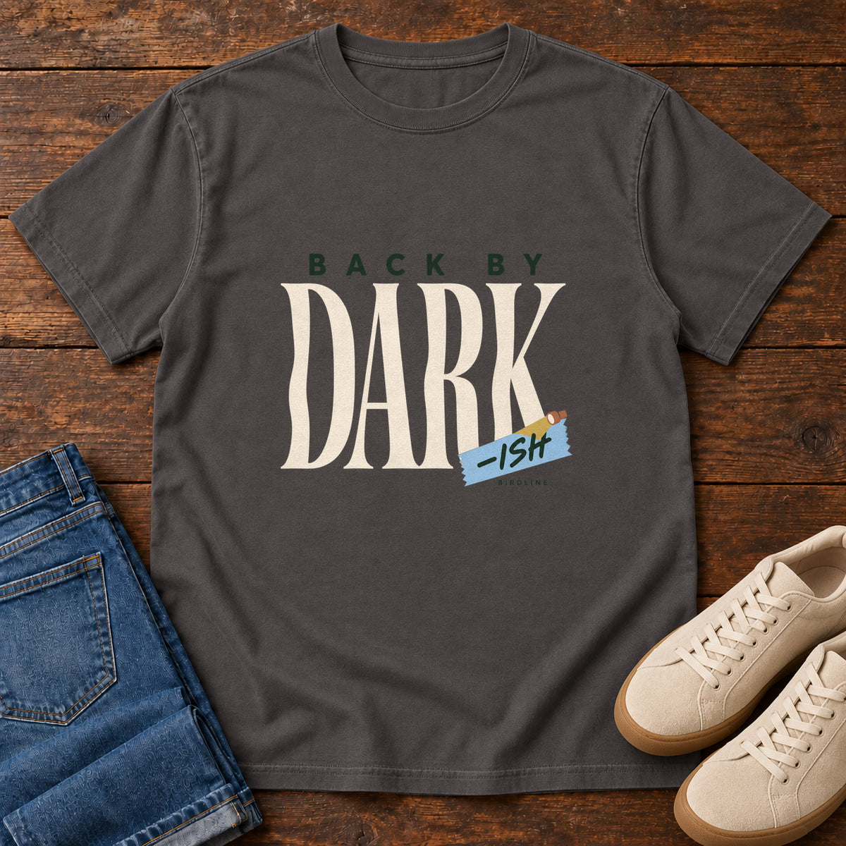 Graphite Back by Dark-ish T-Shirt with tall cream Dark lettering and a small blue -ish label, front flat lay on wood.