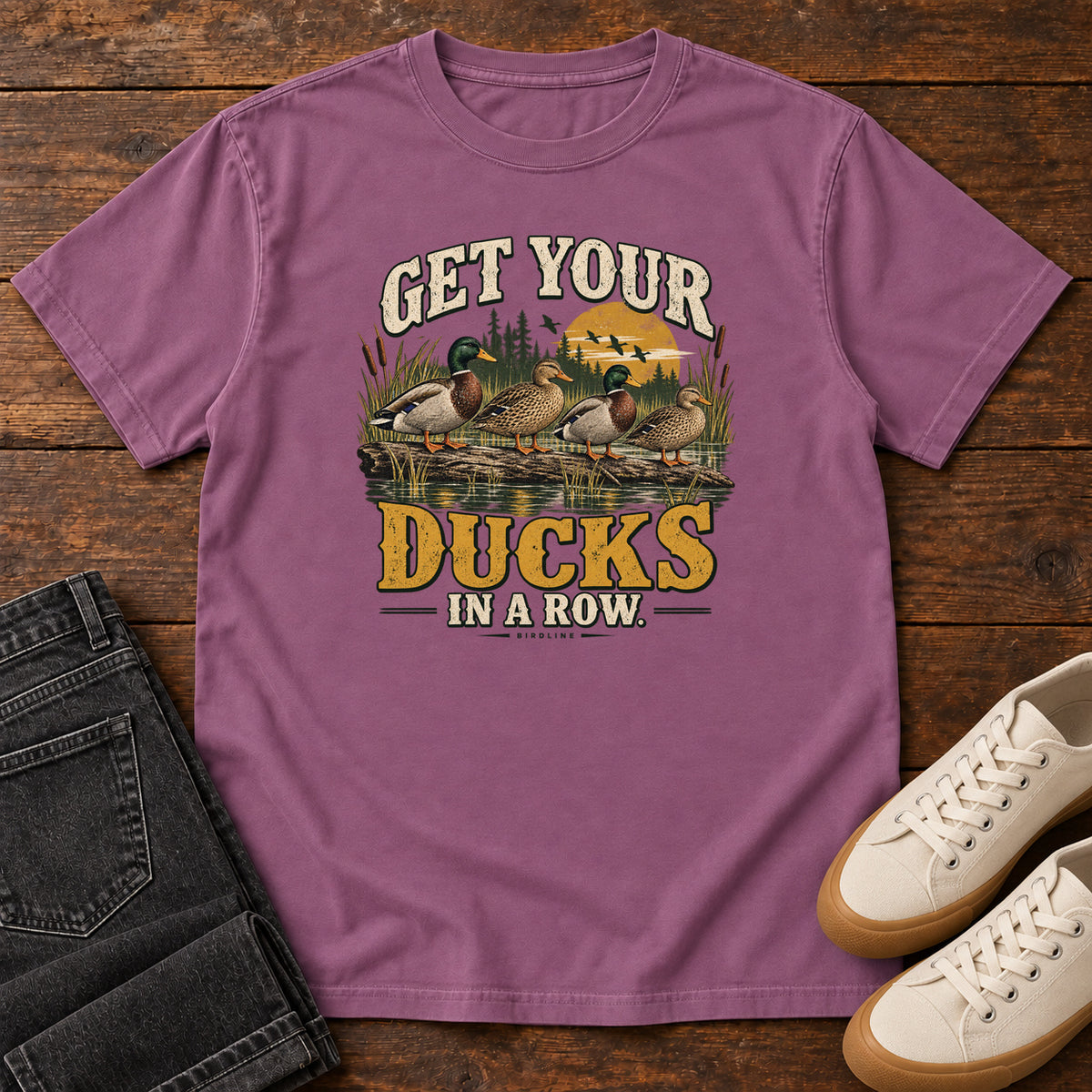 Get Your Ducks in a Row T-Shirt in Berry, front laid flat on wood, featuring a row of ducks among reeds and water beneath cream-and-gold slogan lettering.