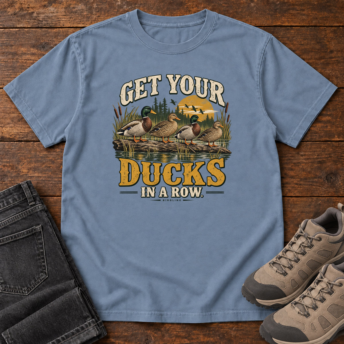 Get Your Ducks in a Row T-Shirt in Blue Jean, front laid flat on wood, featuring a row of ducks among reeds and water beneath cream-and-gold slogan lettering.