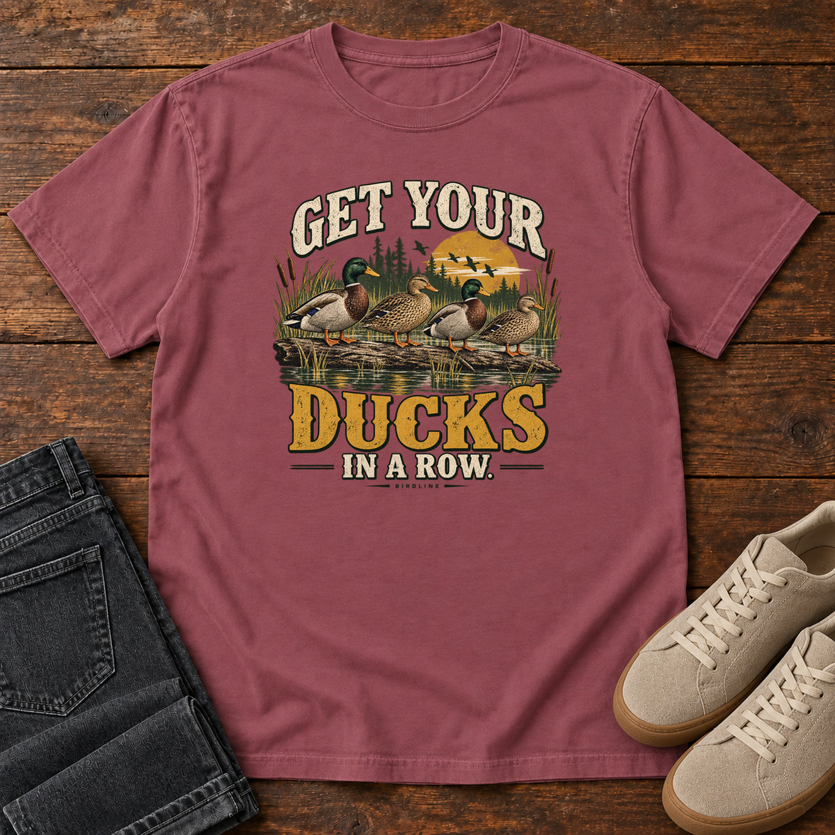 Get Your Ducks in a Row T-Shirt in Brick, front laid flat on wood, featuring a row of ducks among reeds and water beneath cream-and-gold slogan lettering.