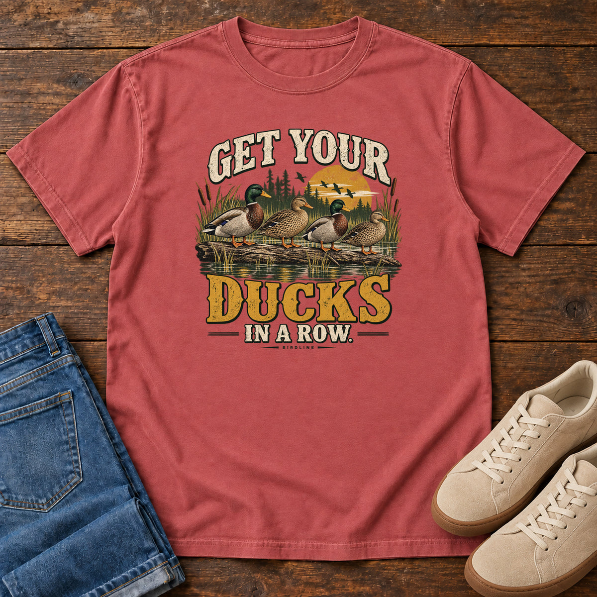 Get Your Ducks in a Row T-Shirt in Crimson, front laid flat on wood, featuring a row of ducks among reeds and water beneath cream-and-gold slogan lettering.