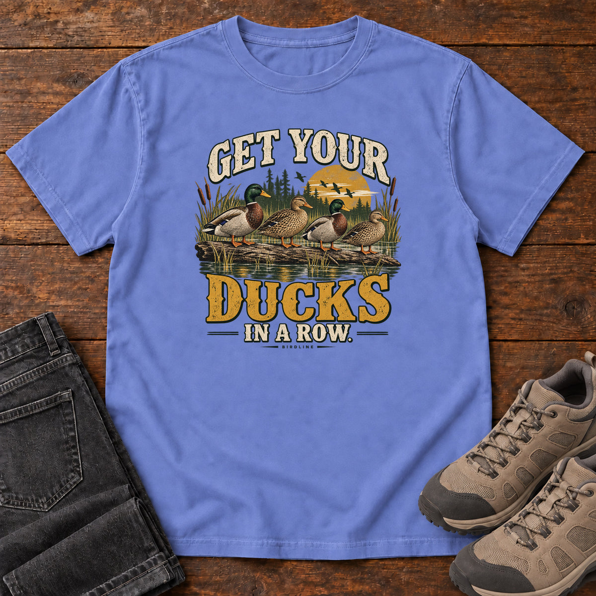 Get Your Ducks in a Row T-Shirt in Flo Blue, front laid flat on wood, featuring a row of ducks among reeds and water beneath cream-and-gold slogan lettering.