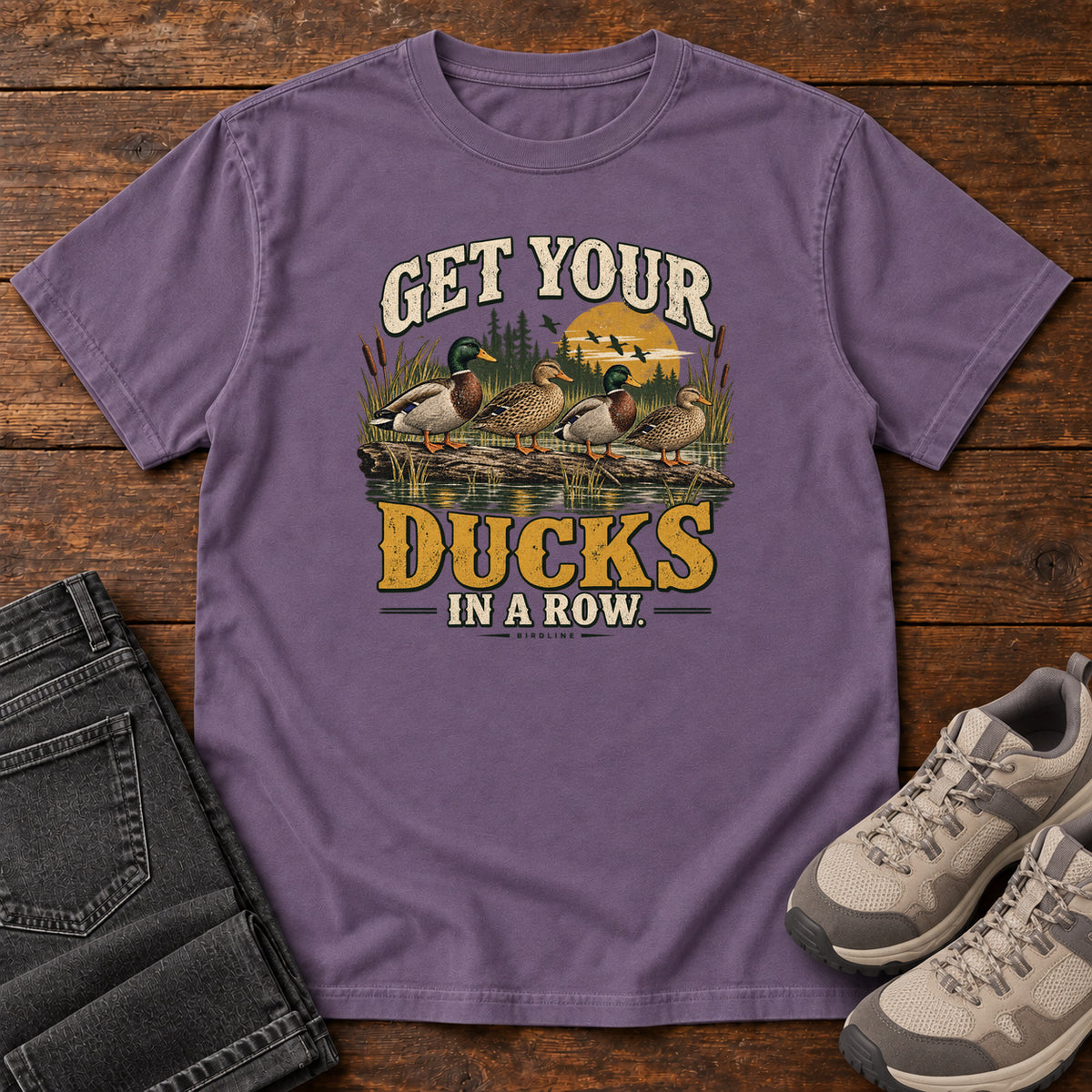 Get Your Ducks in a Row T-Shirt in Grape, front laid flat on wood, featuring a row of ducks among reeds and water beneath cream-and-gold slogan lettering.