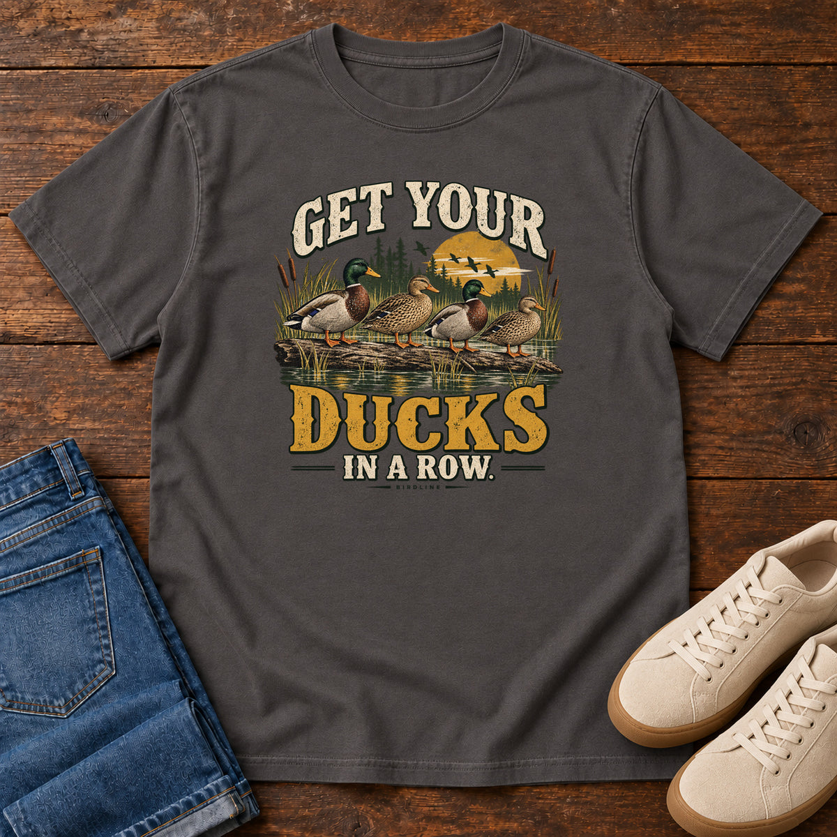 Get Your Ducks in a Row T-Shirt in Graphite, front laid flat on wood, featuring a row of ducks among reeds and water beneath cream-and-gold slogan lettering.