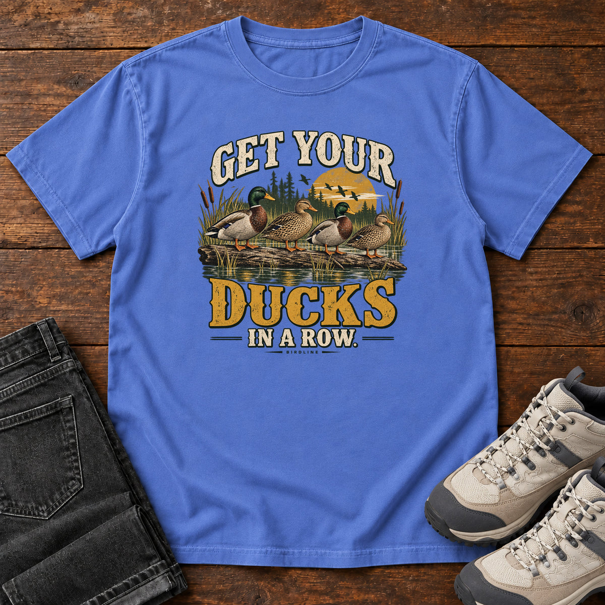 Get Your Ducks in a Row T-Shirt in Mystic Blue, front laid flat on wood, featuring a row of ducks among reeds and water beneath cream-and-gold slogan lettering.