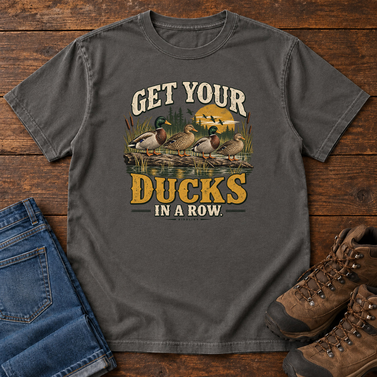 Get Your Ducks in a Row T-Shirt in Pepper, front laid flat on wood, featuring a row of ducks among reeds and water beneath cream-and-gold slogan lettering.