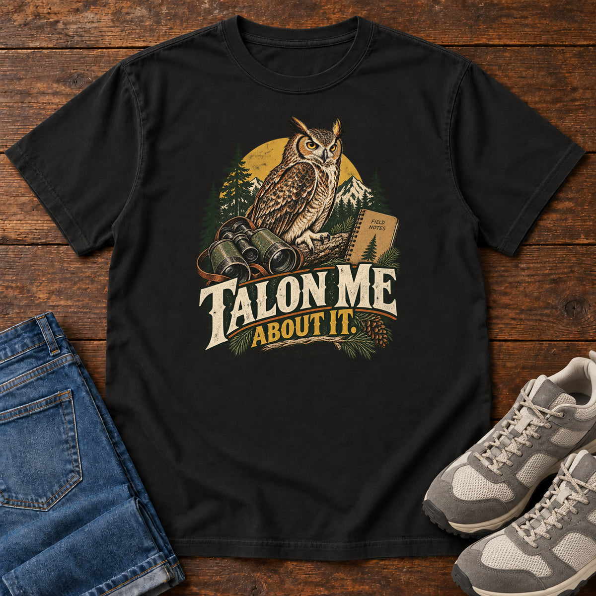 Black Talon Me About It T-Shirt with an owl, binoculars and a field notebook against a forest backdrop, front flat lay on wood.
