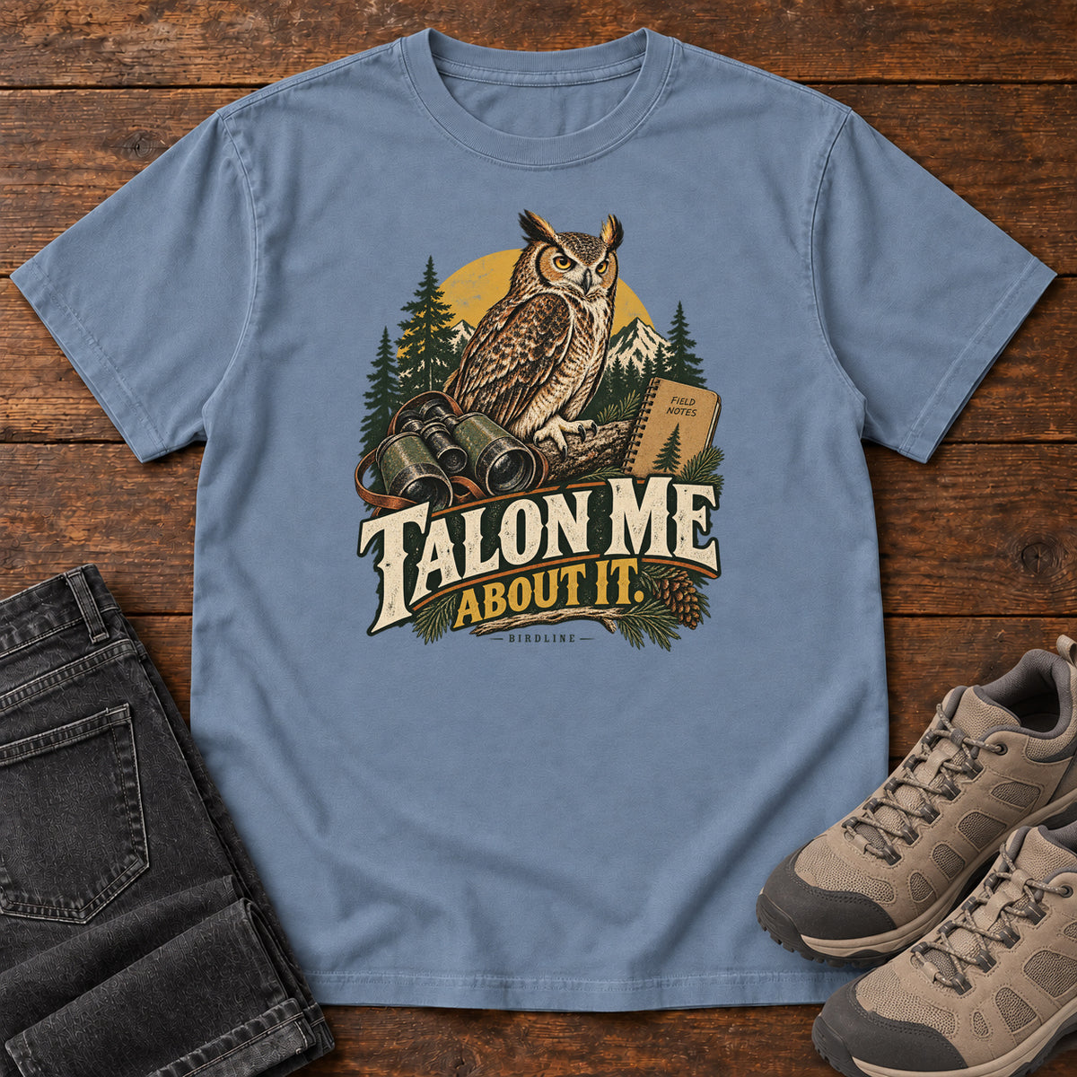 Blue Jean Talon Me About It T-Shirt with an owl, binoculars and a field notebook against a forest backdrop, front flat lay on wood.