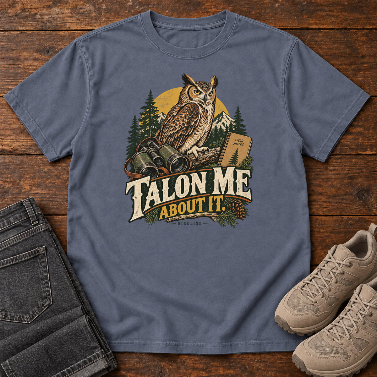 Denim Talon Me About It T-Shirt with an owl, binoculars and a field notebook against a forest backdrop, front flat lay on wood.