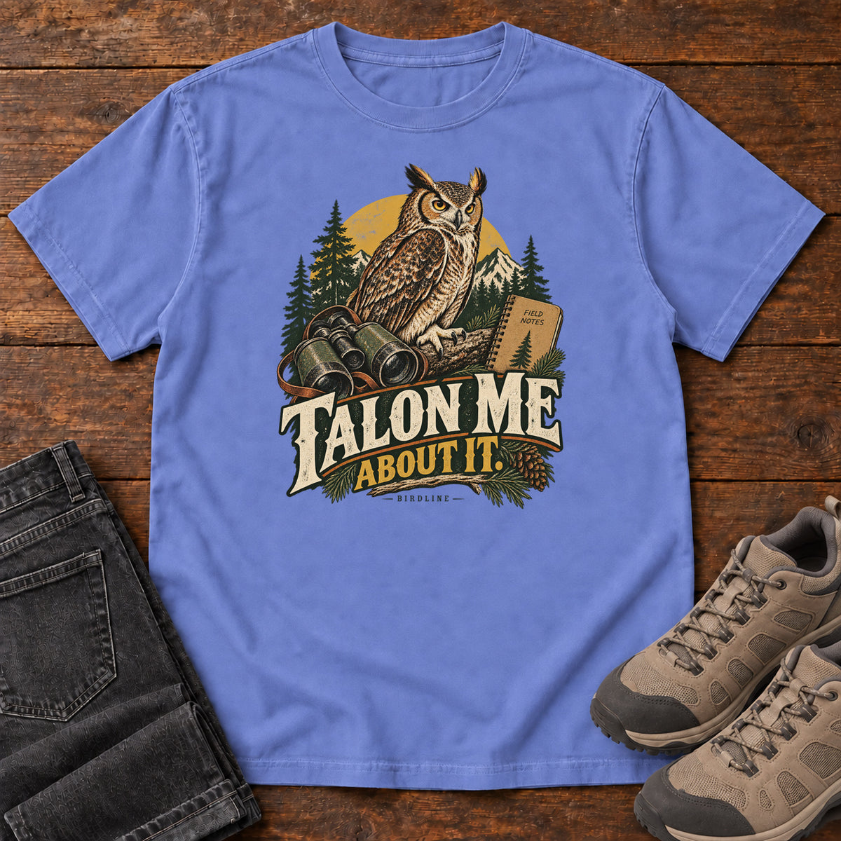 Flo Blue Talon Me About It T-Shirt with an owl, binoculars and a field notebook against a forest backdrop, front flat lay on wood.