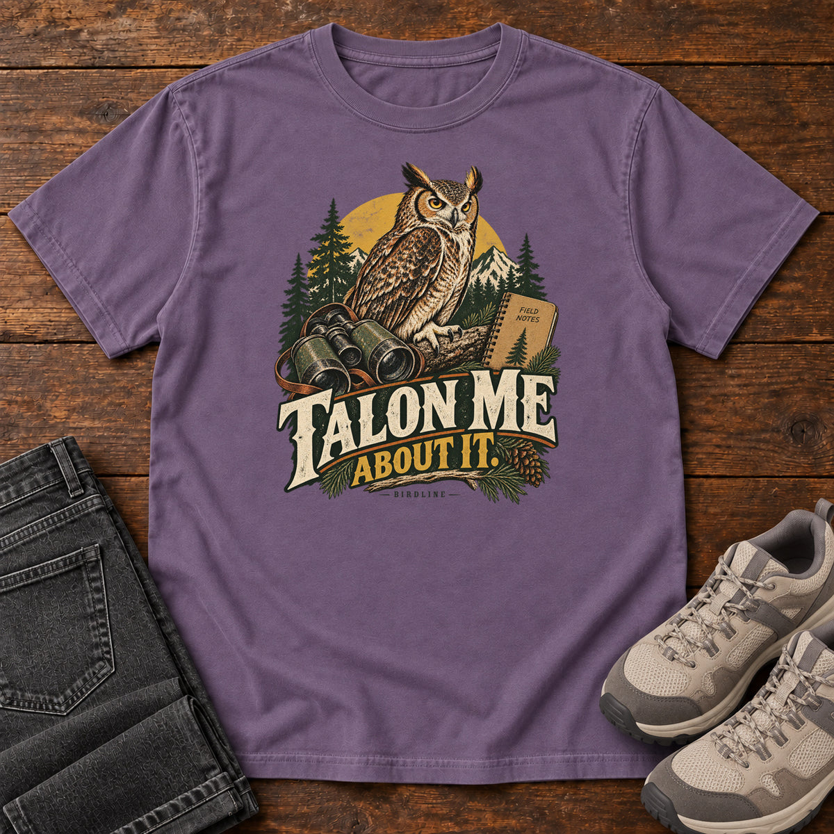 Grape Talon Me About It T-Shirt with an owl, binoculars and a field notebook against a forest backdrop, front flat lay on wood.