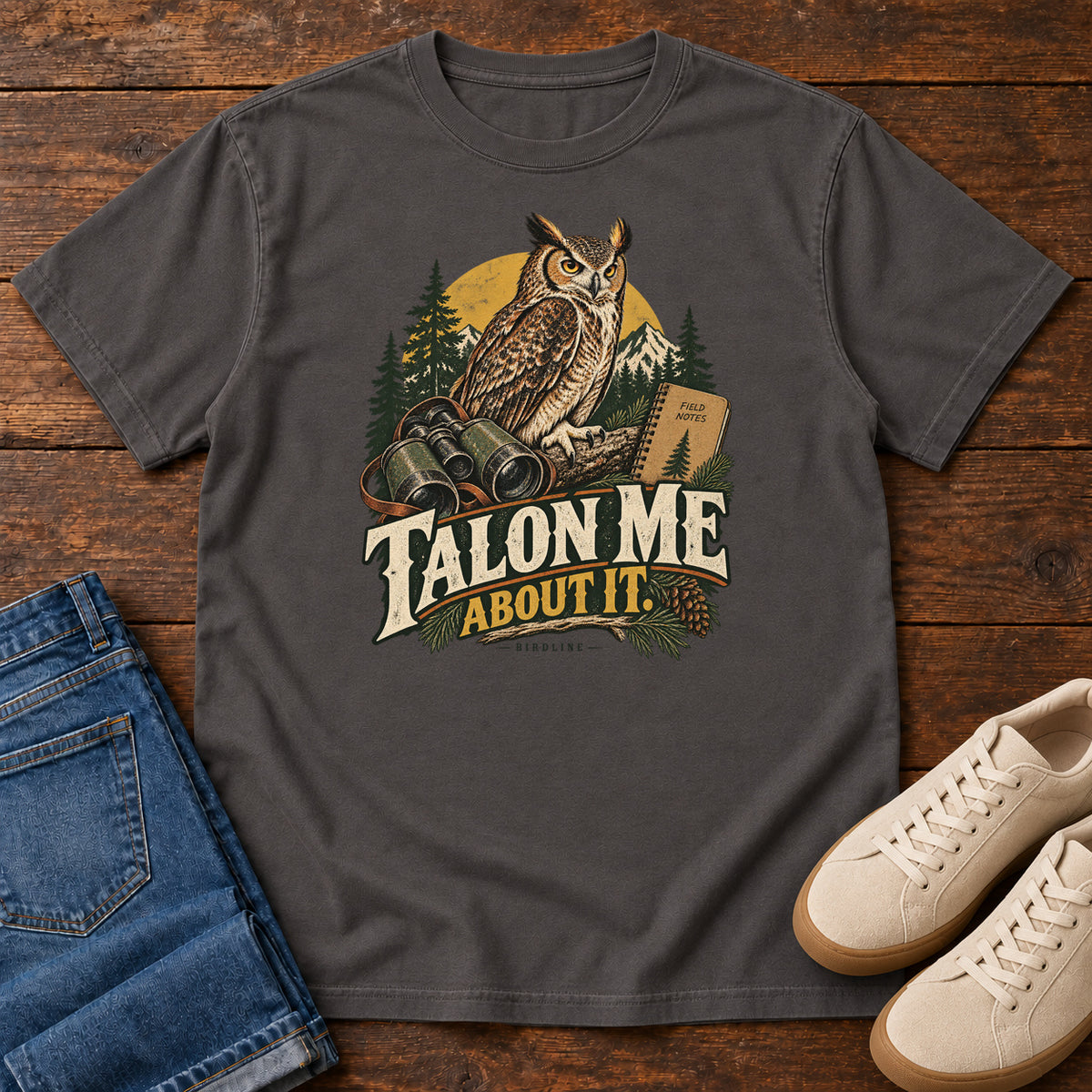 Graphite Talon Me About It T-Shirt with an owl, binoculars and a field notebook against a forest backdrop, front flat lay on wood.
