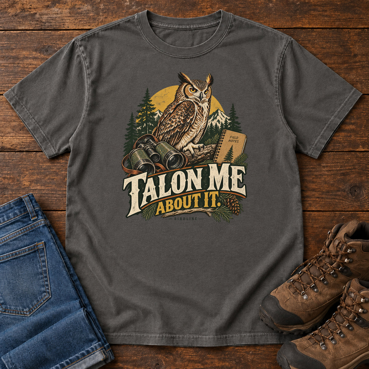 Pepper Talon Me About It T-Shirt with an owl, binoculars and a field notebook against a forest backdrop, front flat lay on wood.