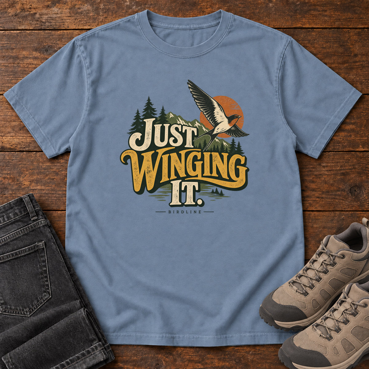 Blue Jean Just Winging It T-Shirt with a flying bird, evergreens, an orange sun and cream and gold lettering; front flat lay on wood.
