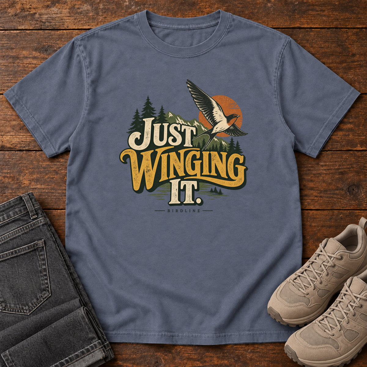 Denim Just Winging It T-Shirt with a flying bird, evergreens, an orange sun and cream and gold lettering; front flat lay on wood.