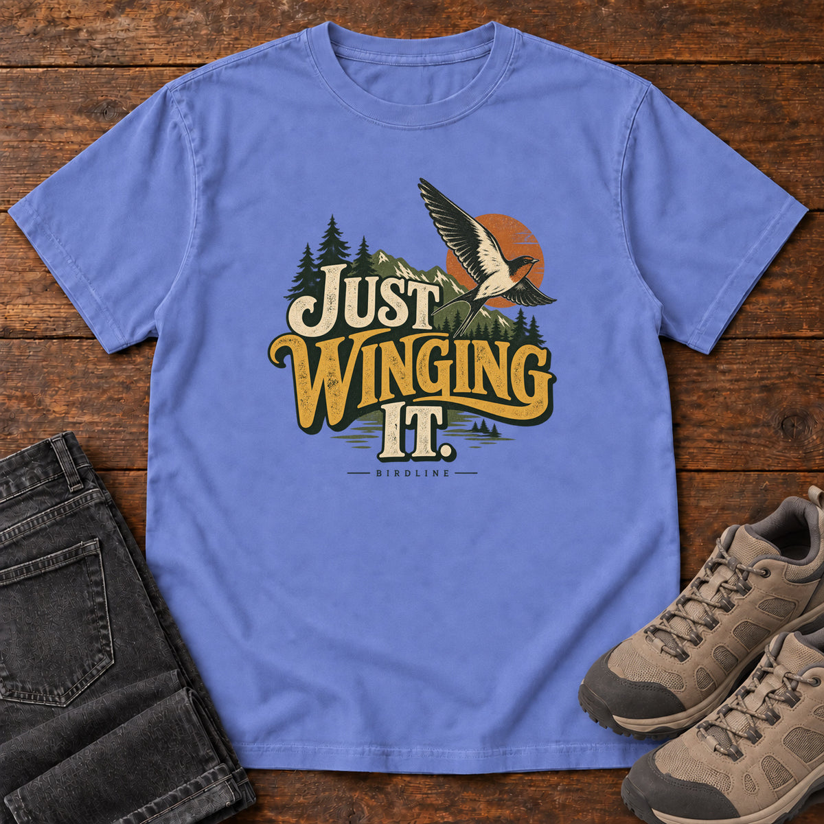 Flo Blue Just Winging It T-Shirt with a flying bird, evergreens, an orange sun and cream and gold lettering; front flat lay on wood.