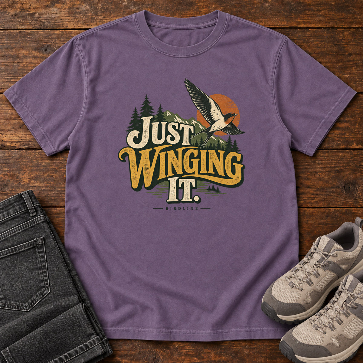 Grape Just Winging It T-Shirt with a flying bird, evergreens, an orange sun and cream and gold lettering; front flat lay on wood.