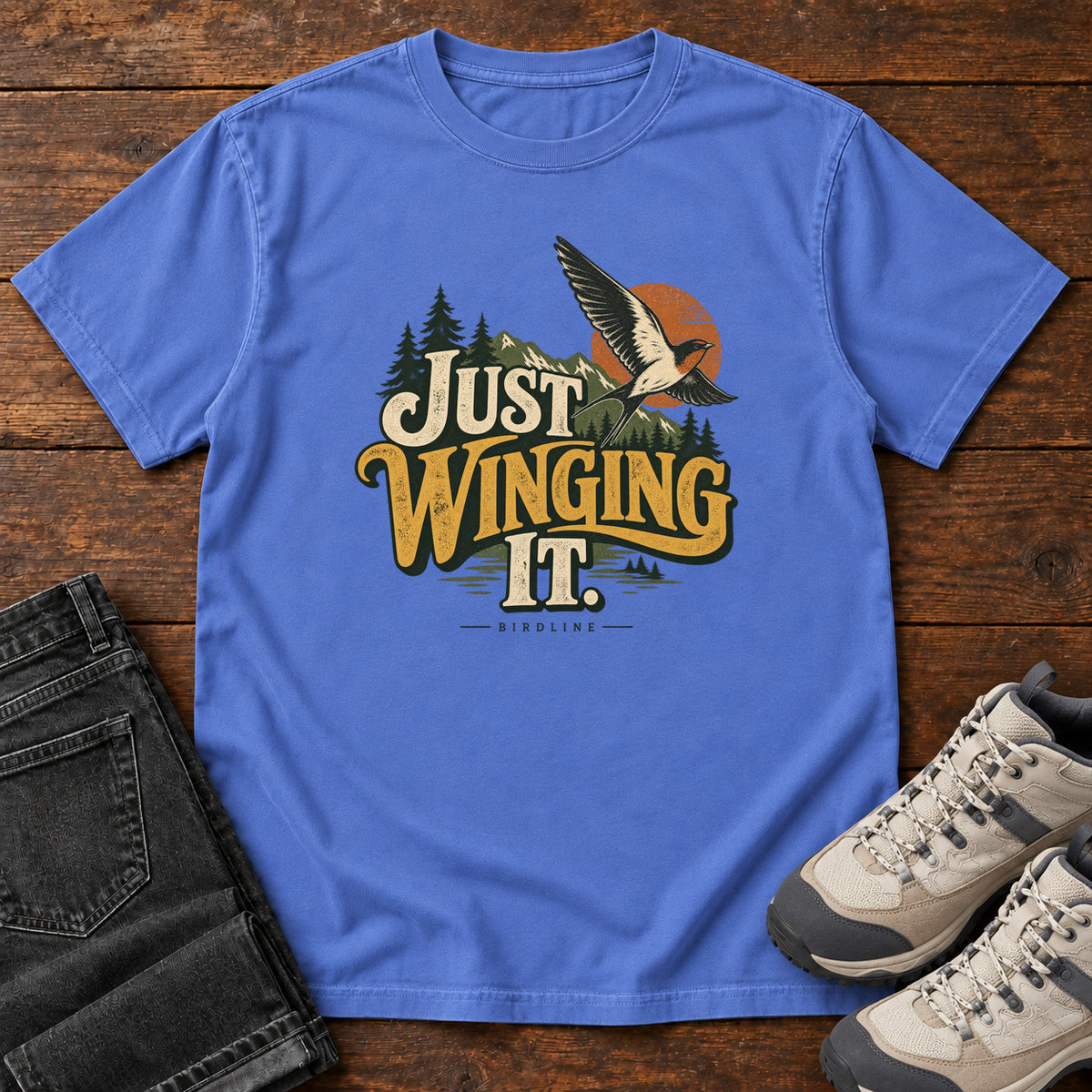 Mystic Blue Just Winging It T-Shirt with a flying bird, evergreens, an orange sun and cream and gold lettering; front flat lay on wood.