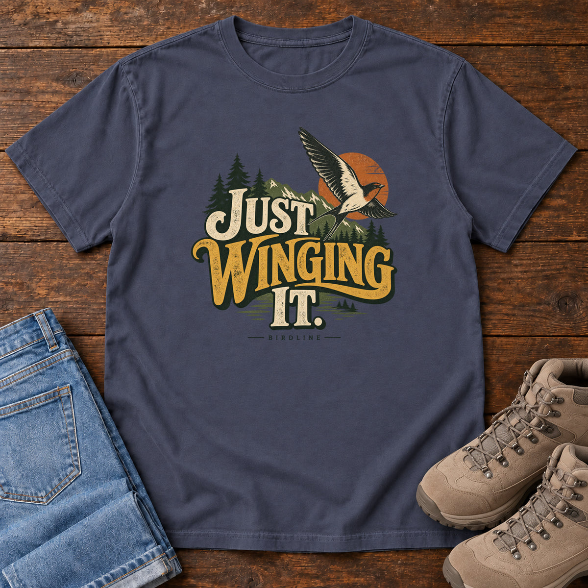 Navy Just Winging It T-Shirt with a flying bird, evergreens, an orange sun and cream and gold lettering; front flat lay on wood.
