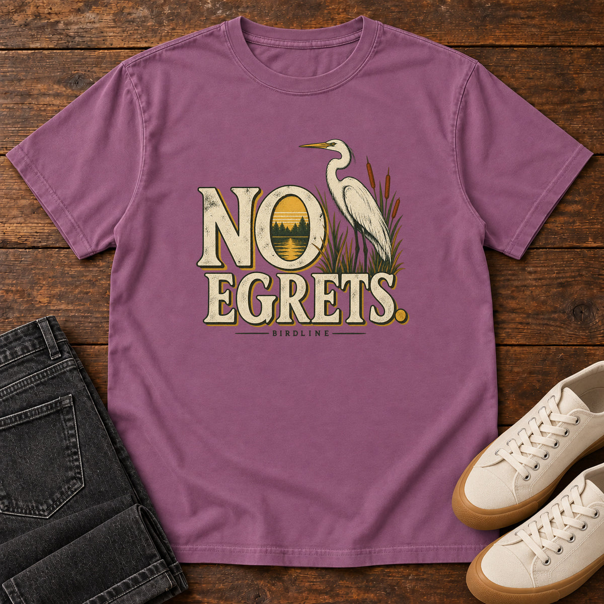 Berry No Egrets. T-Shirt with a white long-necked wading bird beside reeds and lettering framing a sunset; front flat lay on wood.