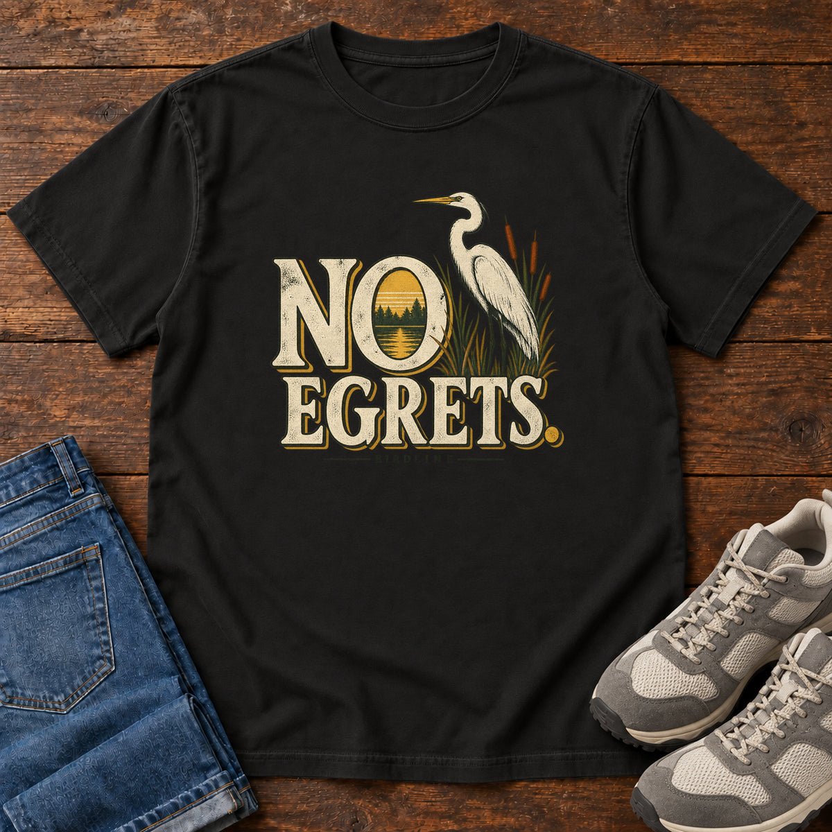 Black No Egrets. T-Shirt with a white long-necked wading bird beside reeds and lettering framing a sunset; front flat lay on wood.