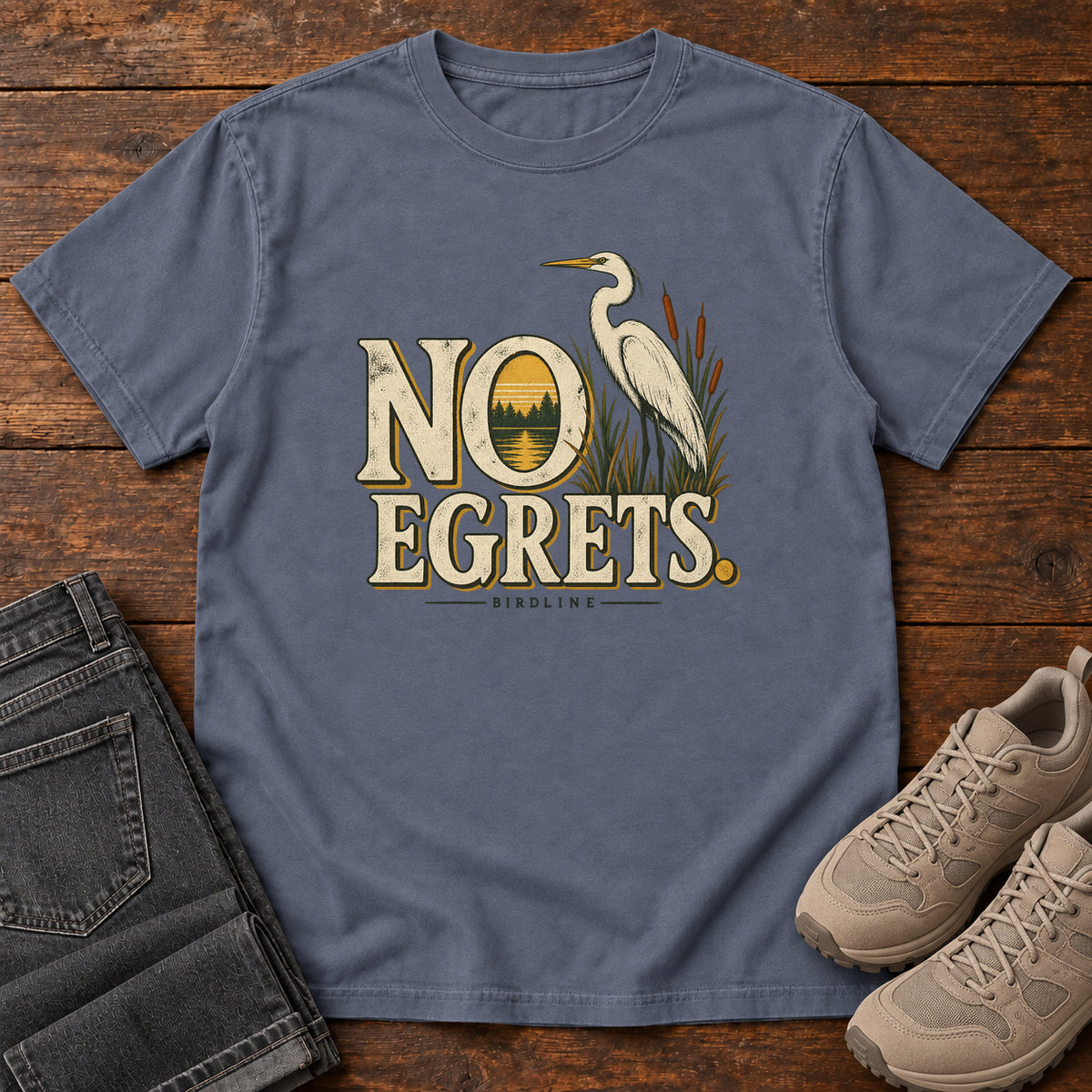 Denim No Egrets. T-Shirt with a white long-necked wading bird beside reeds and lettering framing a sunset; front flat lay on wood.