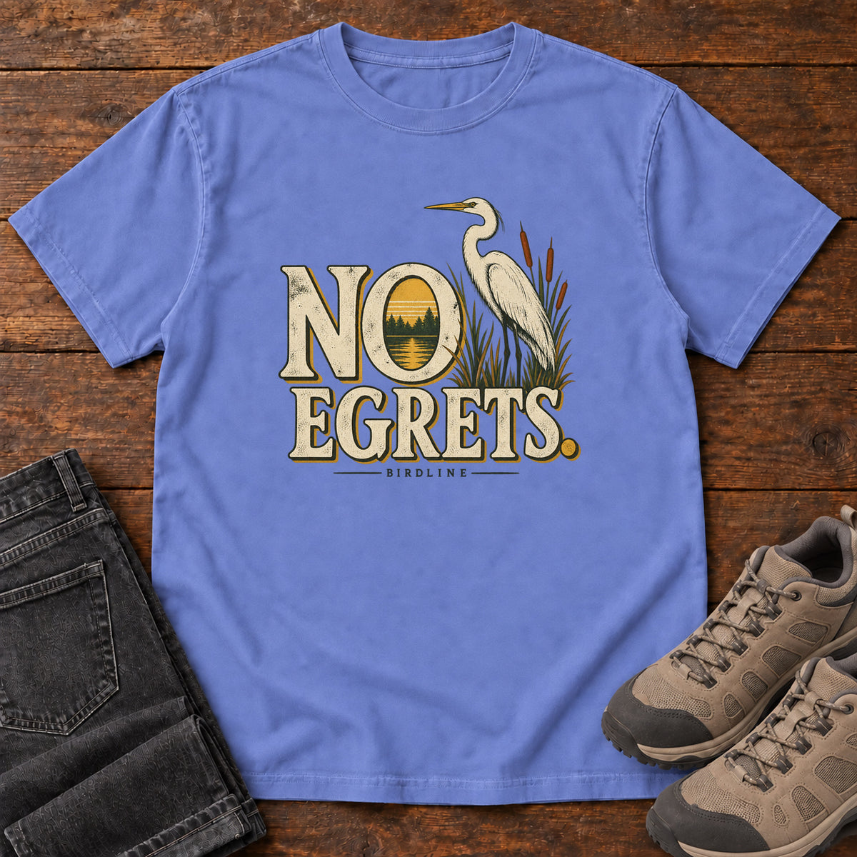 Flo Blue No Egrets. T-Shirt with a white long-necked wading bird beside reeds and lettering framing a sunset; front flat lay on wood.