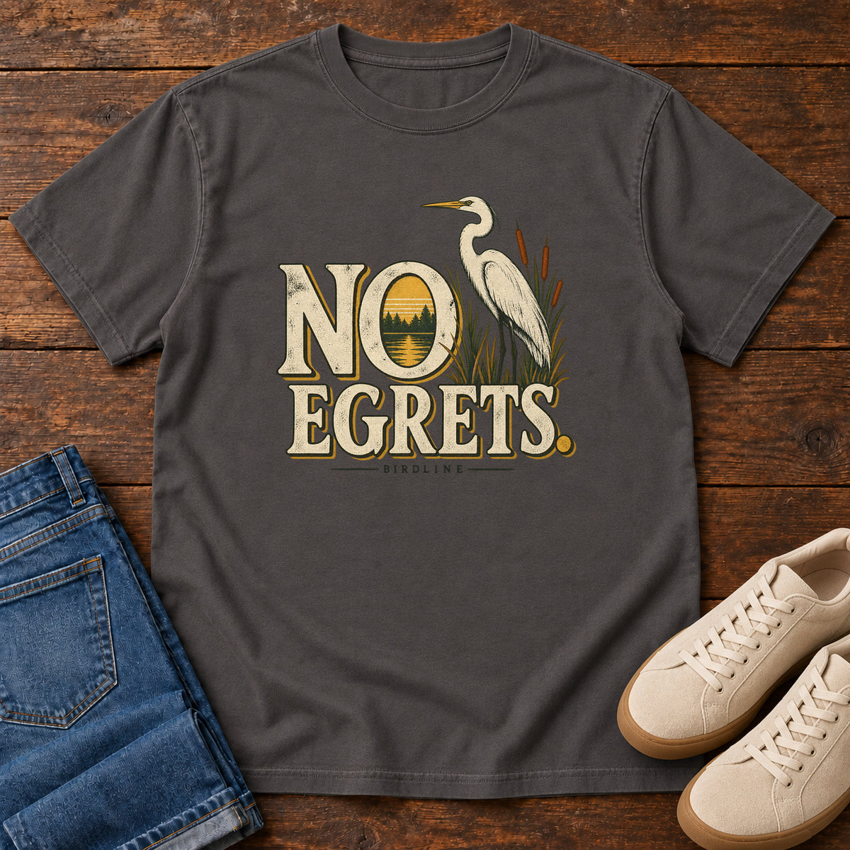 Graphite No Egrets. T-Shirt with a white long-necked wading bird beside reeds and lettering framing a sunset; front flat lay on wood.