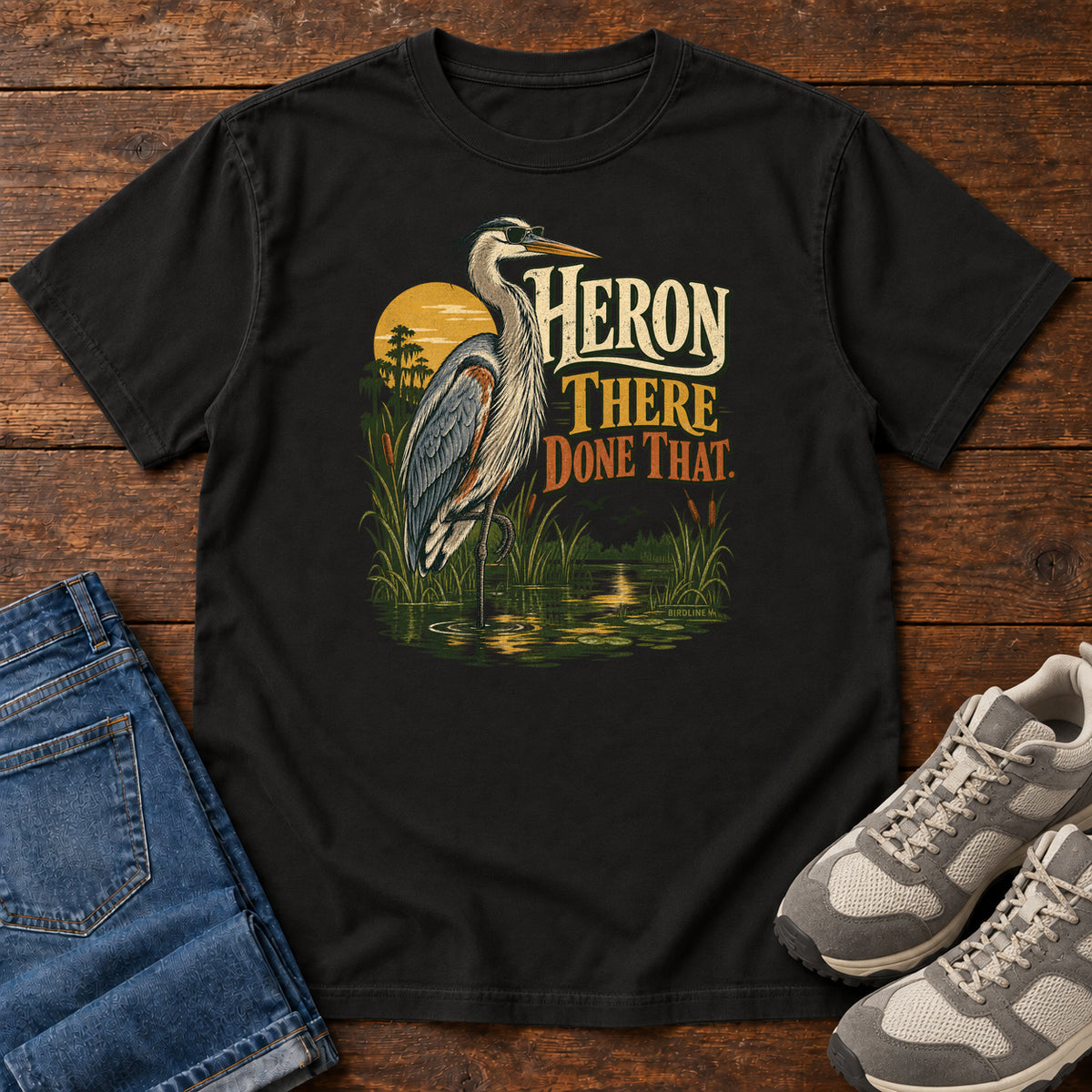 Black Heron There Done That T-Shirt with a long-legged gray bird standing in marsh water beside sunset and cream and orange lettering, front flat lay on wood.