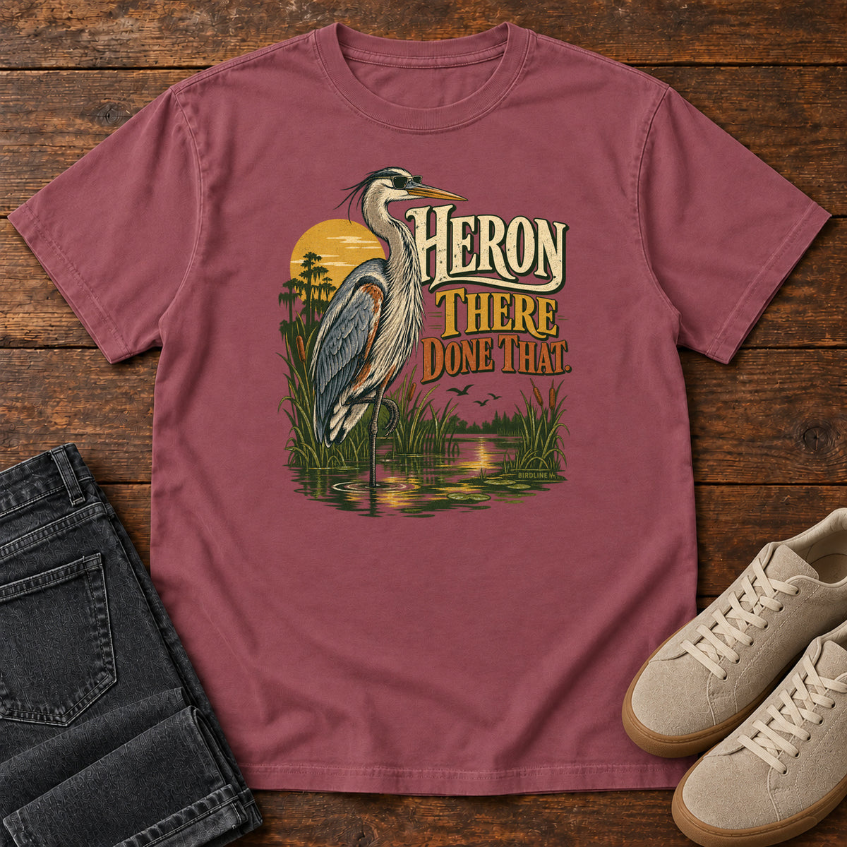 Brick Heron There Done That T-Shirt with a long-legged gray bird standing in marsh water beside sunset and cream and orange lettering, front flat lay on wood.