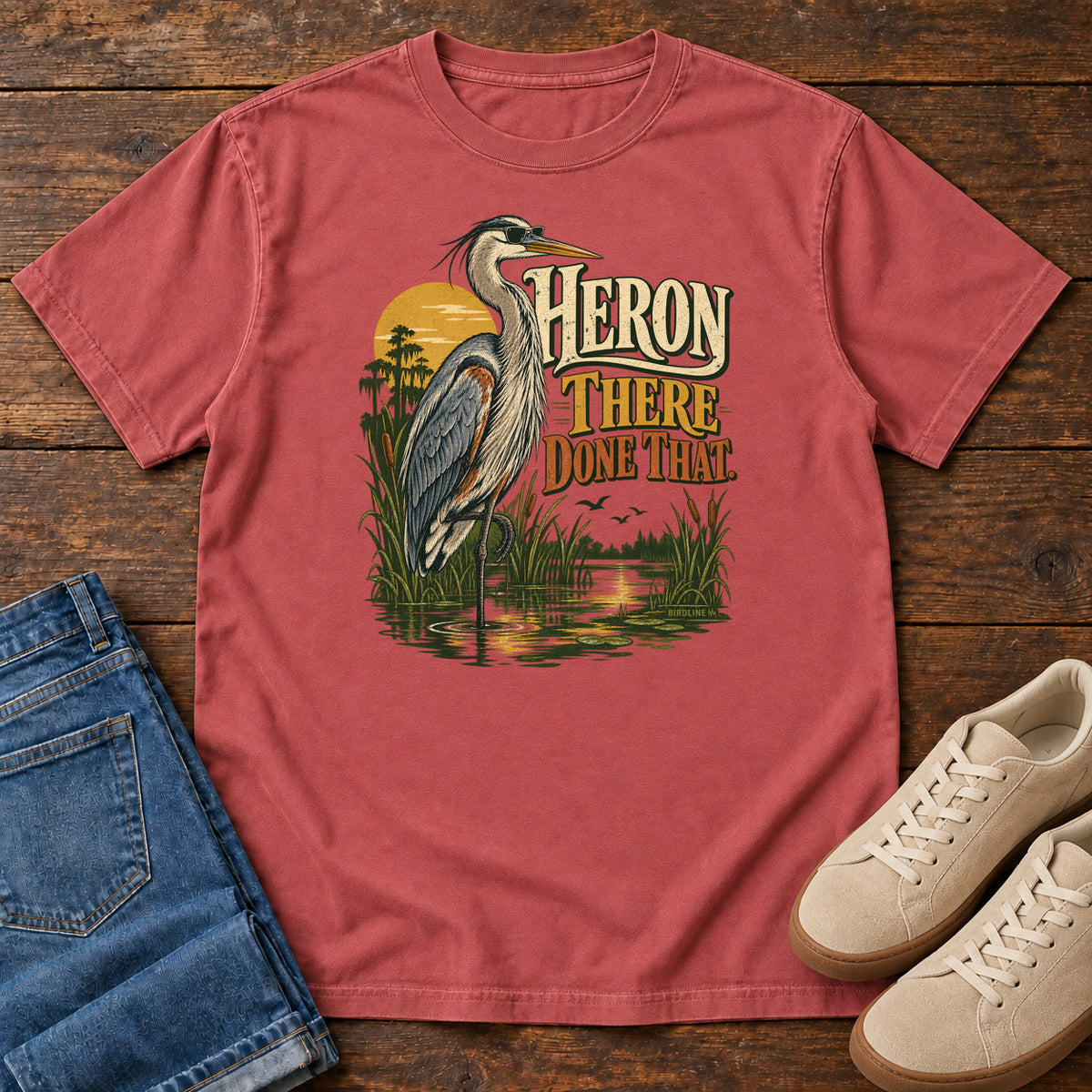 Crimson Heron There Done That T-Shirt with a long-legged gray bird standing in marsh water beside sunset and cream and orange lettering, front flat lay on wood.