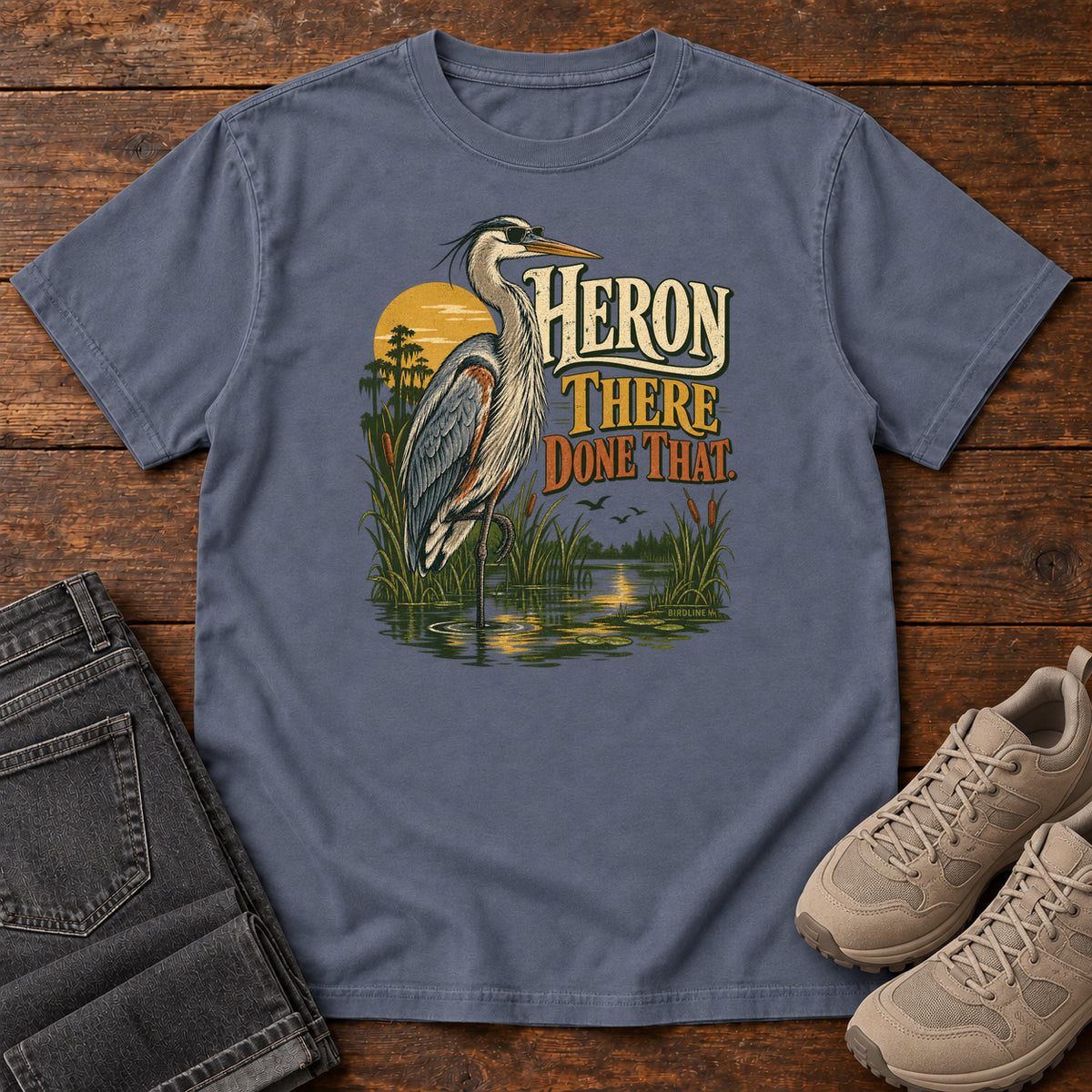 Denim Heron There Done That T-Shirt with a long-legged gray bird standing in marsh water beside sunset and cream and orange lettering, front flat lay on wood.