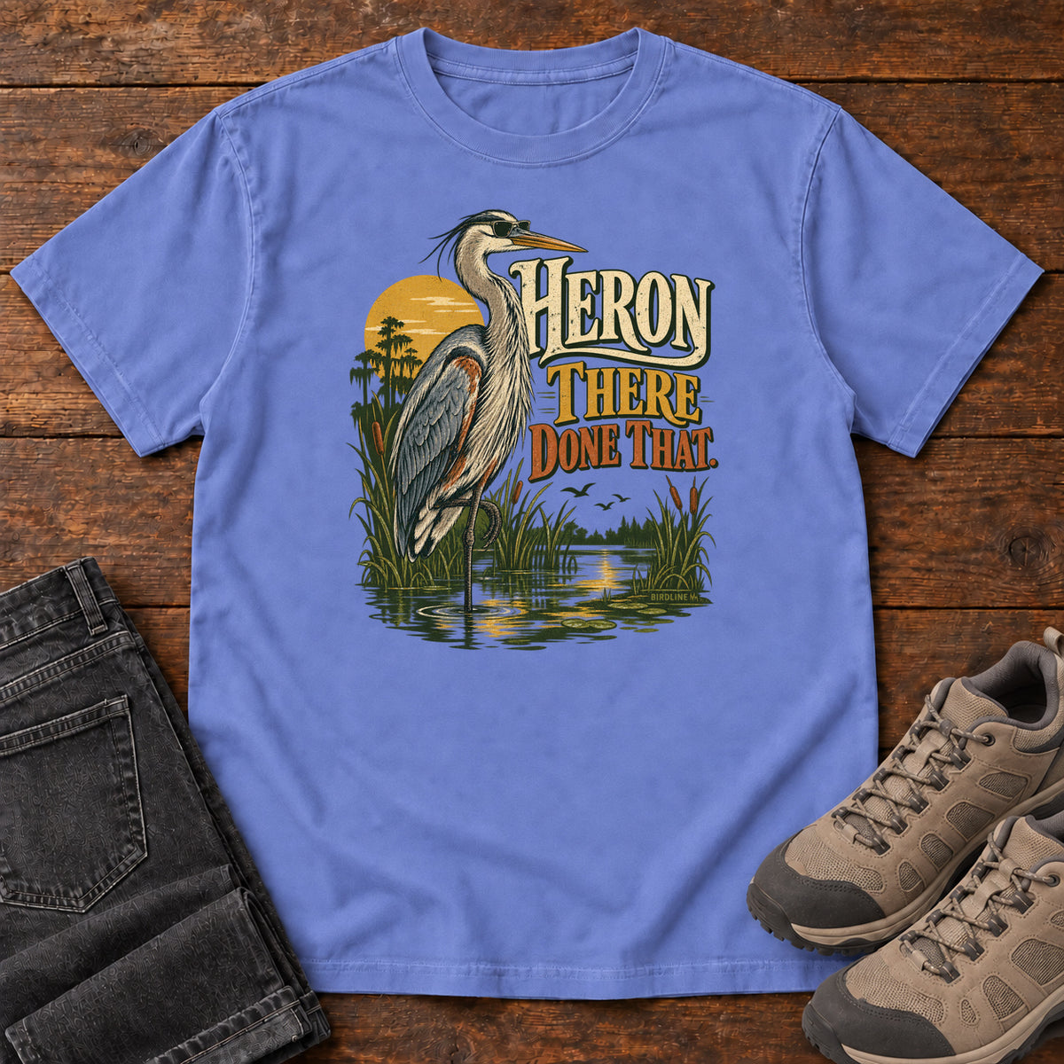 Flo Blue Heron There Done That T-Shirt with a long-legged gray bird standing in marsh water beside sunset and cream and orange lettering, front flat lay on wood.
