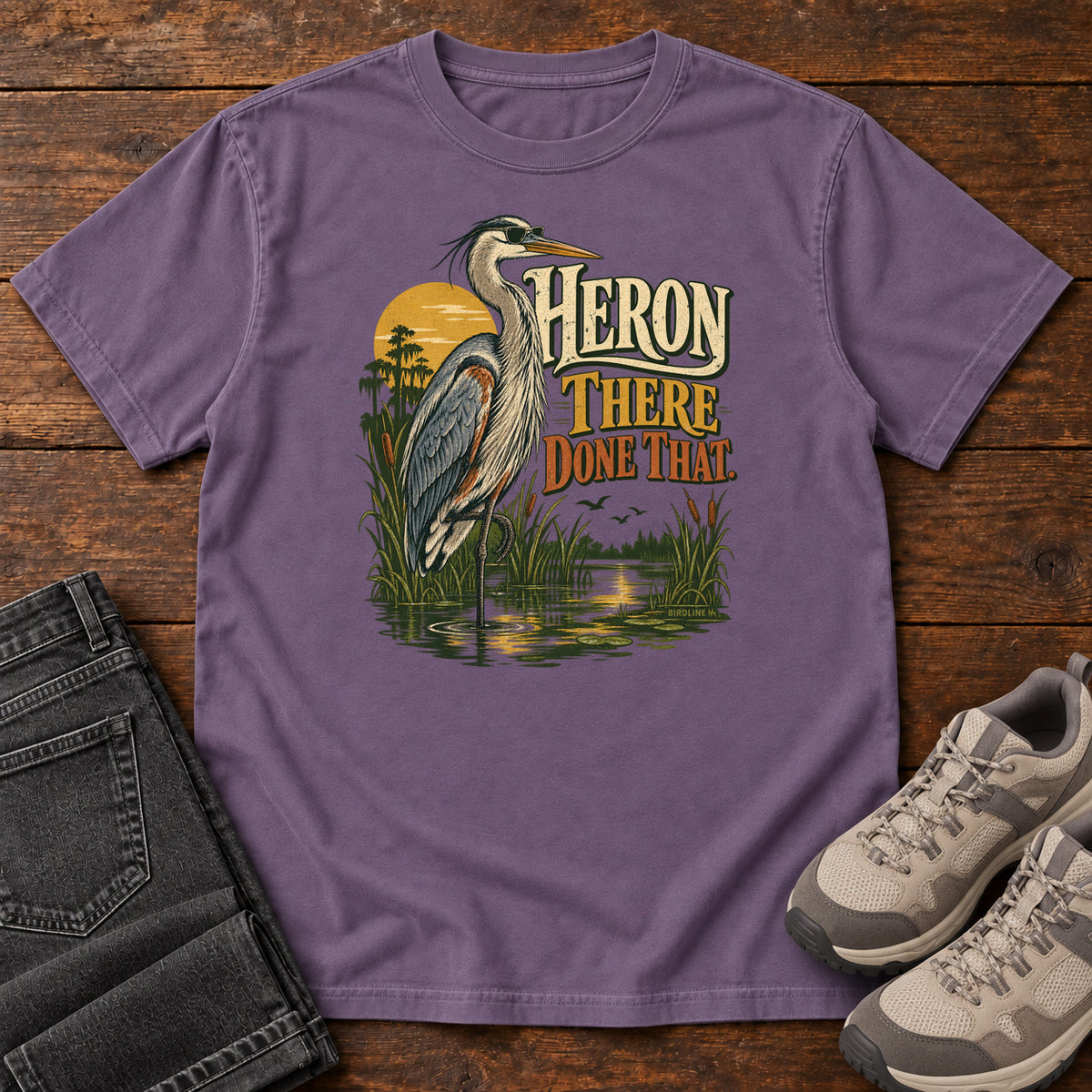 Grape Heron There Done That T-Shirt with a long-legged gray bird standing in marsh water beside sunset and cream and orange lettering, front flat lay on wood.