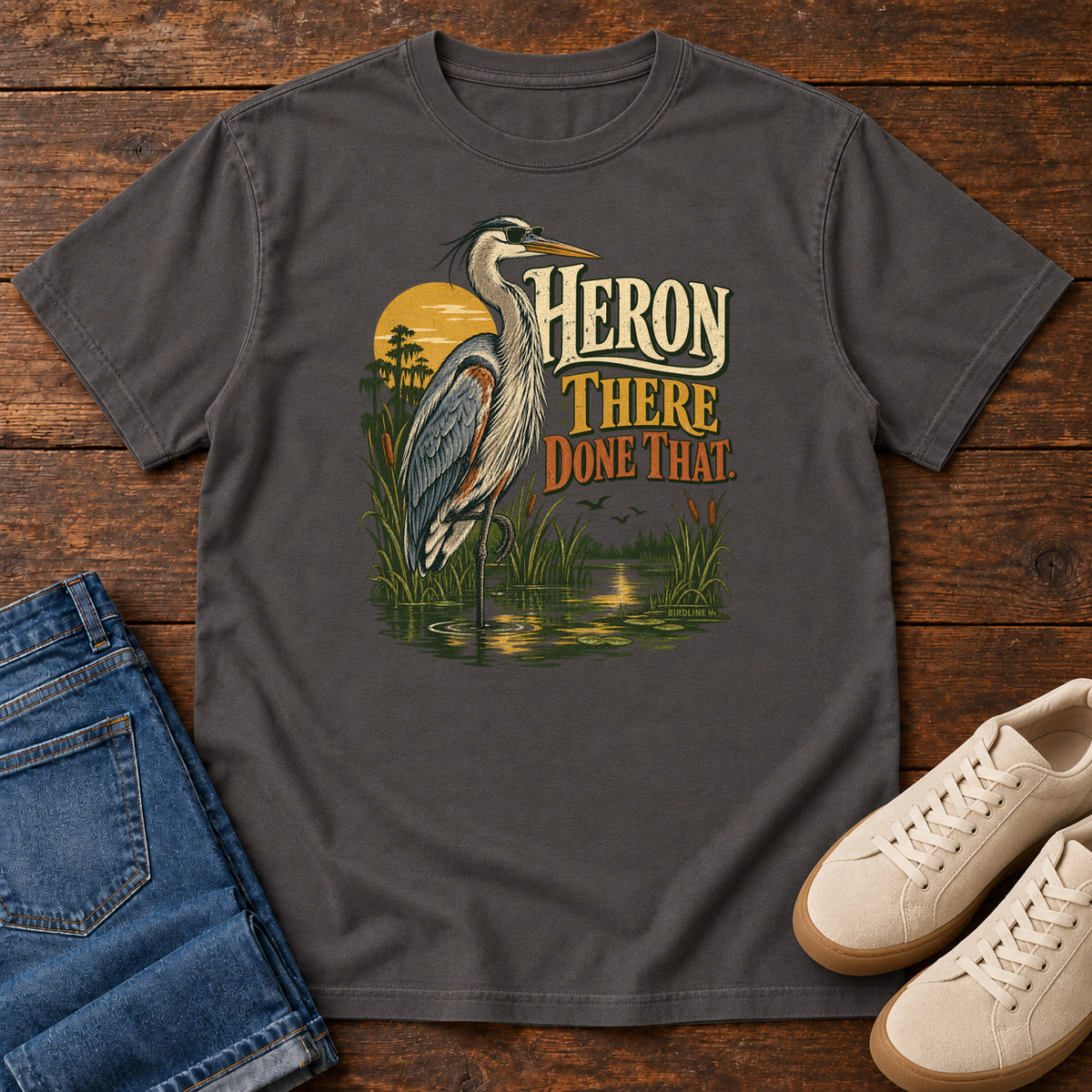 Graphite Heron There Done That T-Shirt with a long-legged gray bird standing in marsh water beside sunset and cream and orange lettering, front flat lay on wood.