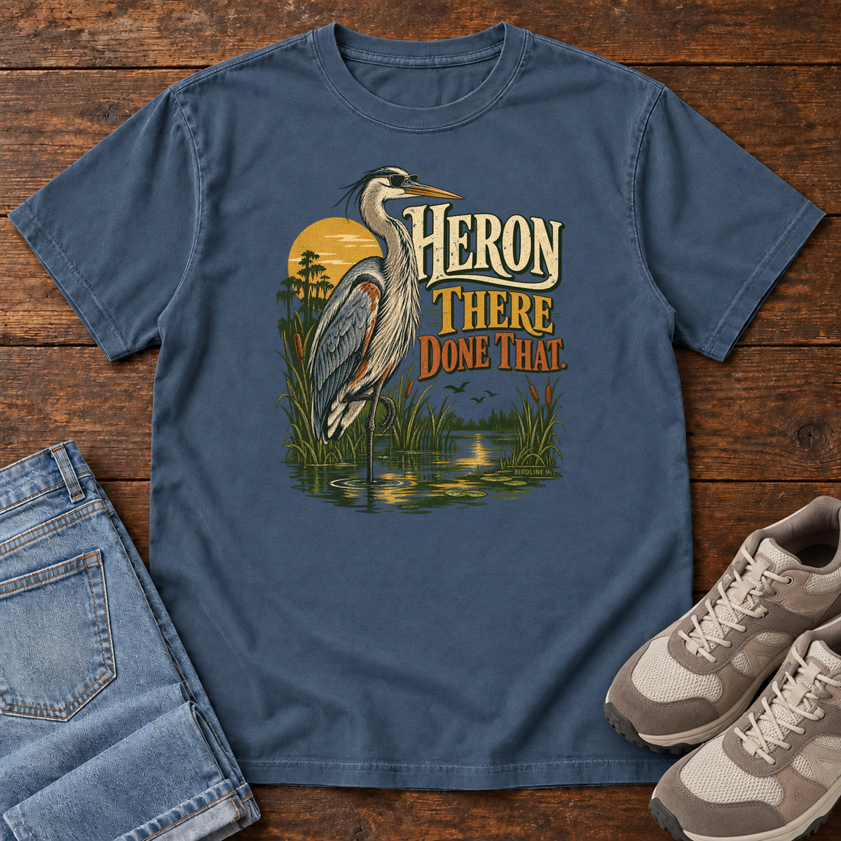 Midnight Heron There Done That T-Shirt with a long-legged gray bird standing in marsh water beside sunset and cream and orange lettering, front flat lay on wood.