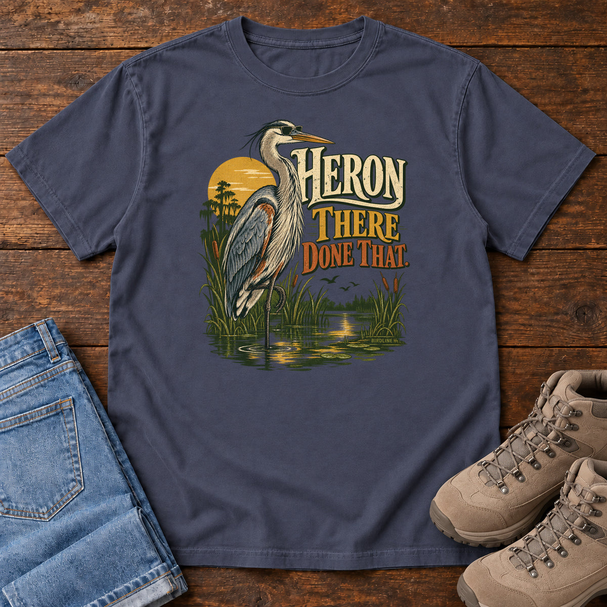 Navy Heron There Done That T-Shirt with a long-legged gray bird standing in marsh water beside sunset and cream and orange lettering, front flat lay on wood.