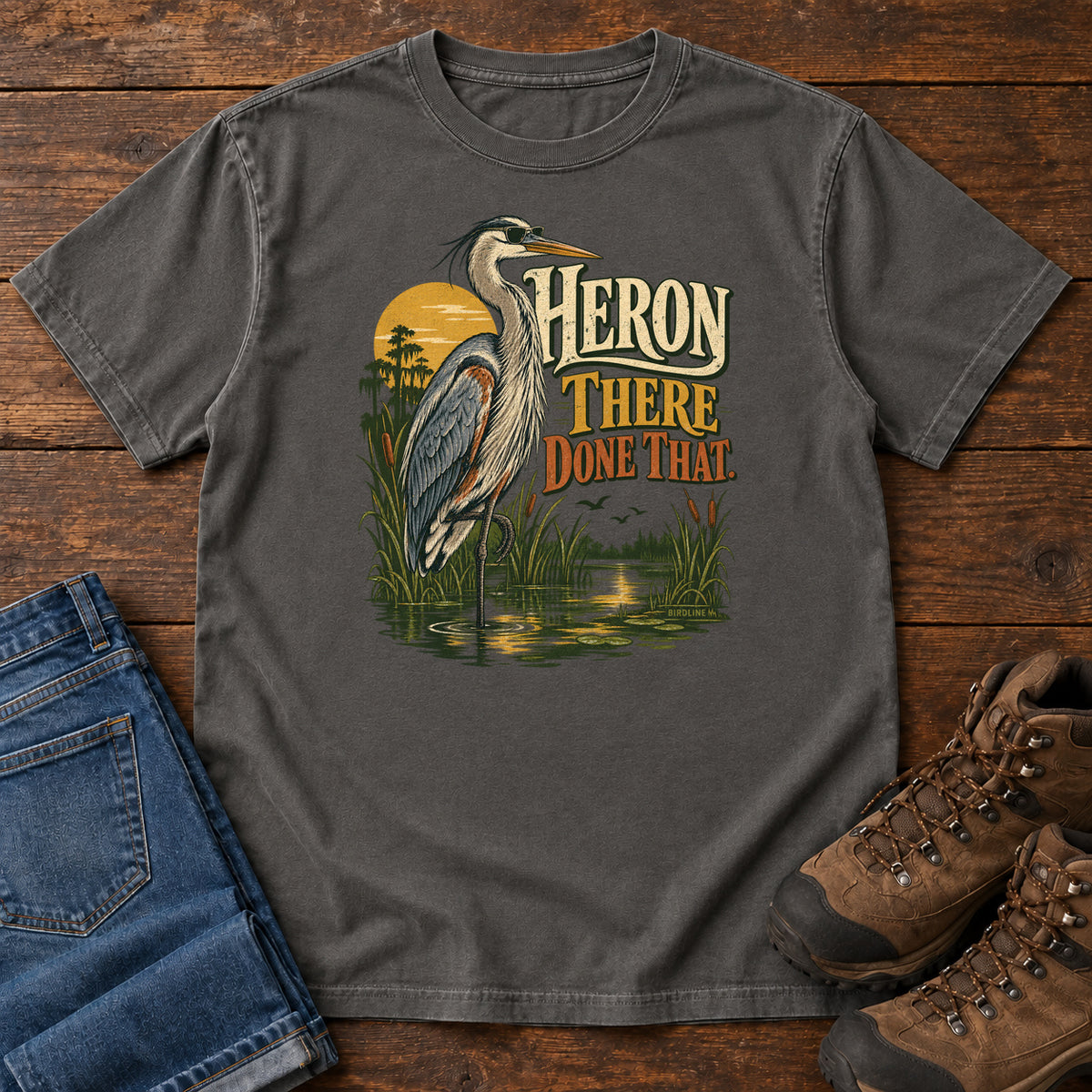 Pepper Heron There Done That T-Shirt with a long-legged gray bird standing in marsh water beside sunset and cream and orange lettering, front flat lay on wood.