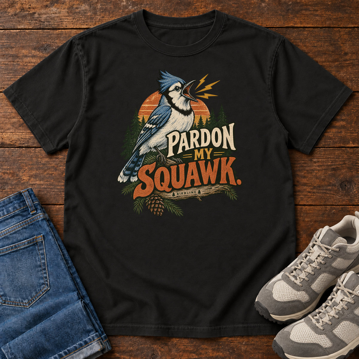 Pardon My Squawk T-Shirt in Black, front laid flat on wood, featuring a blue-and-white crested bird with an open beak, orange sound marks, pine trees and bold lettering.