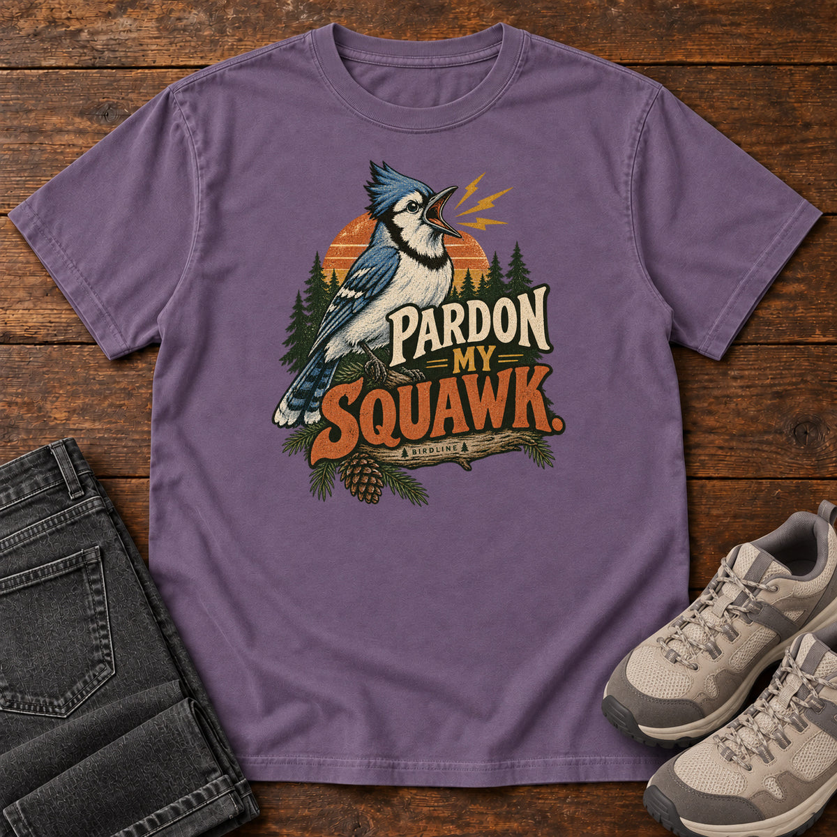 Pardon My Squawk T-Shirt in Grape, front laid flat on wood, featuring a blue-and-white crested bird with an open beak, orange sound marks, pine trees and bold lettering.
