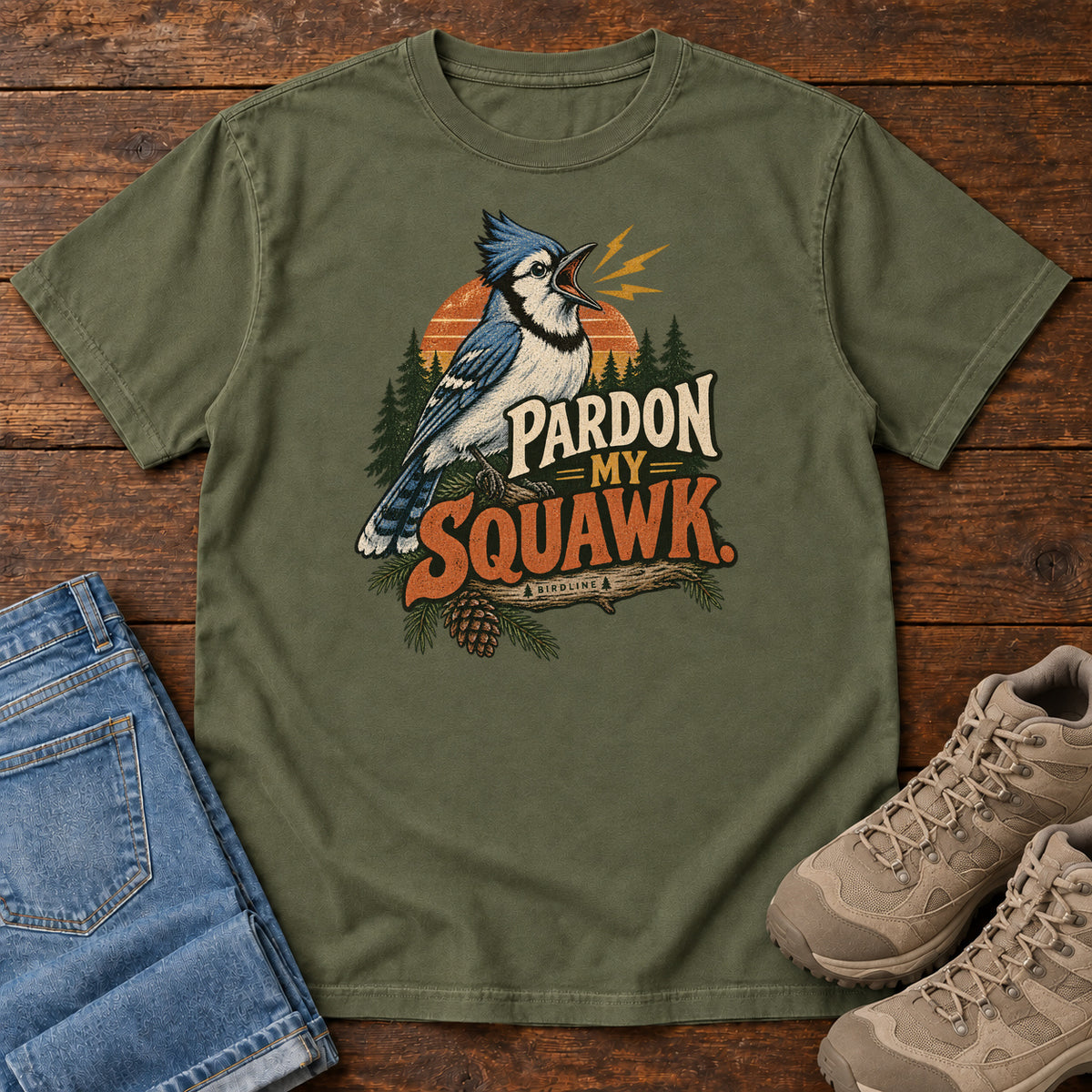Pardon My Squawk T-Shirt in Hemp, front laid flat on wood, featuring a blue-and-white crested bird with an open beak, orange sound marks, pine trees and bold lettering.