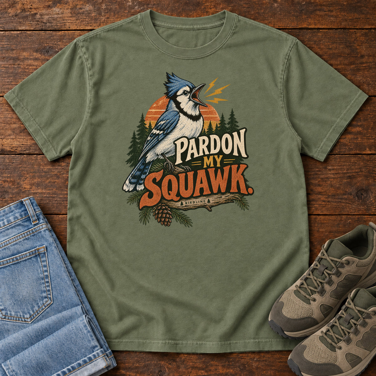 Pardon My Squawk T-Shirt in Moss, front laid flat on wood, featuring a blue-and-white crested bird with an open beak, orange sound marks, pine trees and bold lettering.