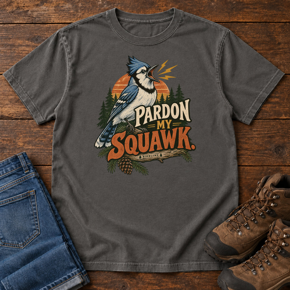 Pardon My Squawk T-Shirt in Pepper, front laid flat on wood, featuring a blue-and-white crested bird with an open beak, orange sound marks, pine trees and bold lettering.