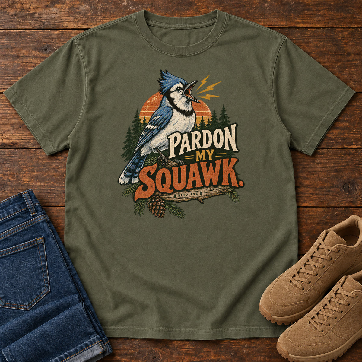 Pardon My Squawk T-Shirt in Sage, front laid flat on wood, featuring a blue-and-white crested bird with an open beak, orange sound marks, pine trees and bold lettering.
